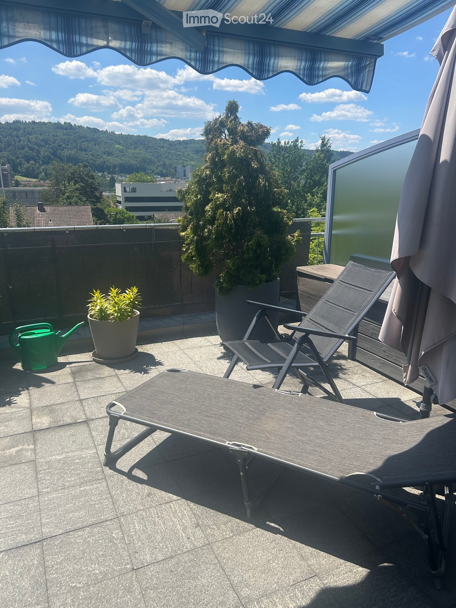 Balcony with lounge chairs, small table, umbrella, potted plants, and a scenic view of the city and hills.