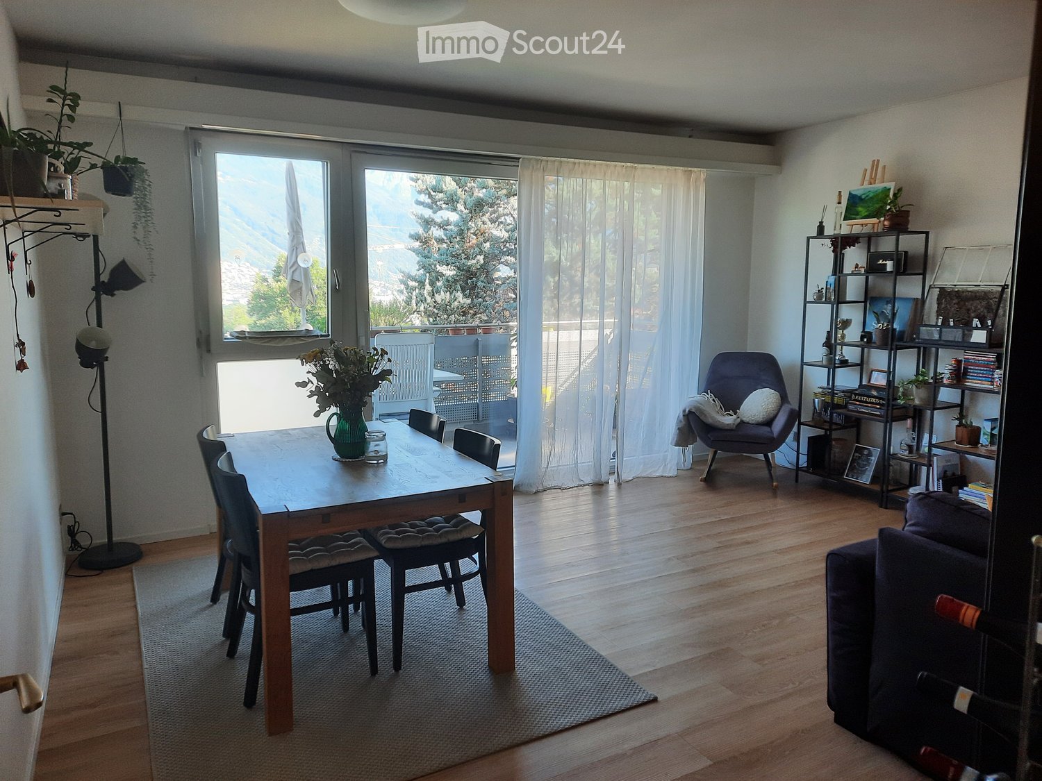 Bright dining area with wooden table, chairs, sofa, shelf, and view of greenery through large windows.
