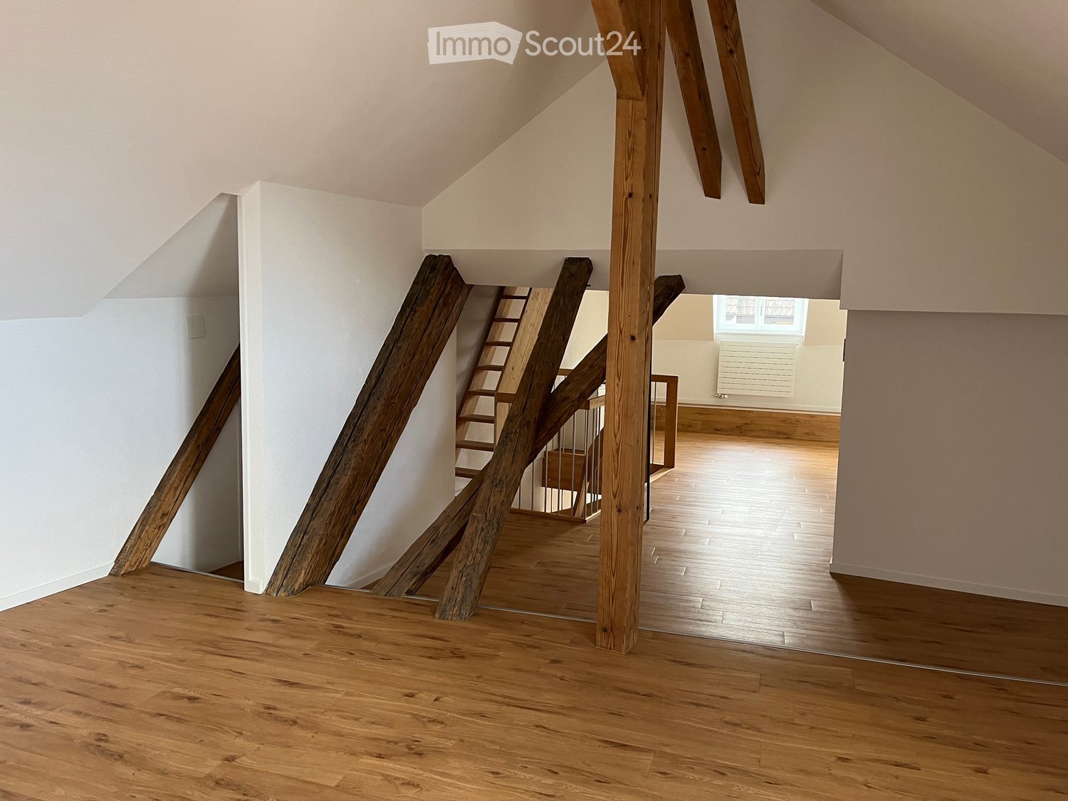 Empty hallway with wooden floors, white walls, and exposed wooden beams. A staircase leads to another room with wooden flooring and white walls.