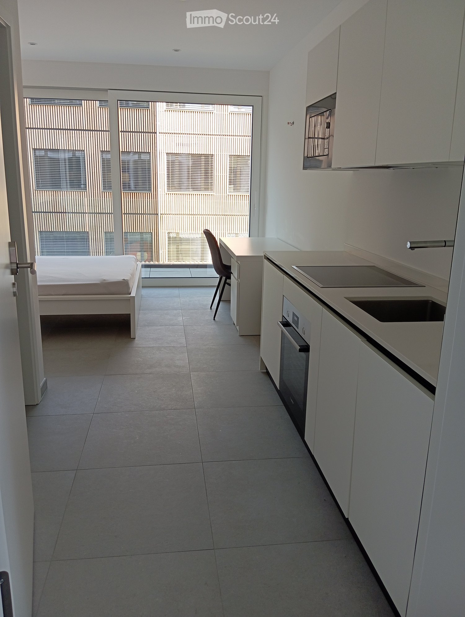 Modern, open-plan studio with a bed, chair, and kitchen area featuring white cabinets, a sink, and an oven. Large windows provide natural light.