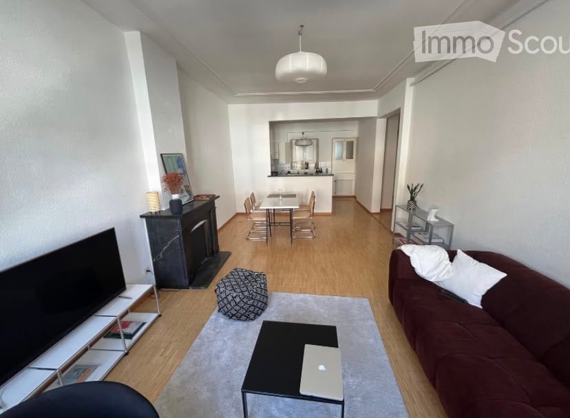 Modern living room with a brown sofa, coffee table, laptop, ottoman, fireplace, and dining area with chairs and a table. The room features light wooden flooring and white walls.