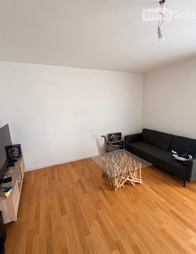 Living room with light wooden flooring, white walls, a black sofa, a unique glass coffee table with a wooden base, a TV stand with a TV, and a small cat sitting on the floor.