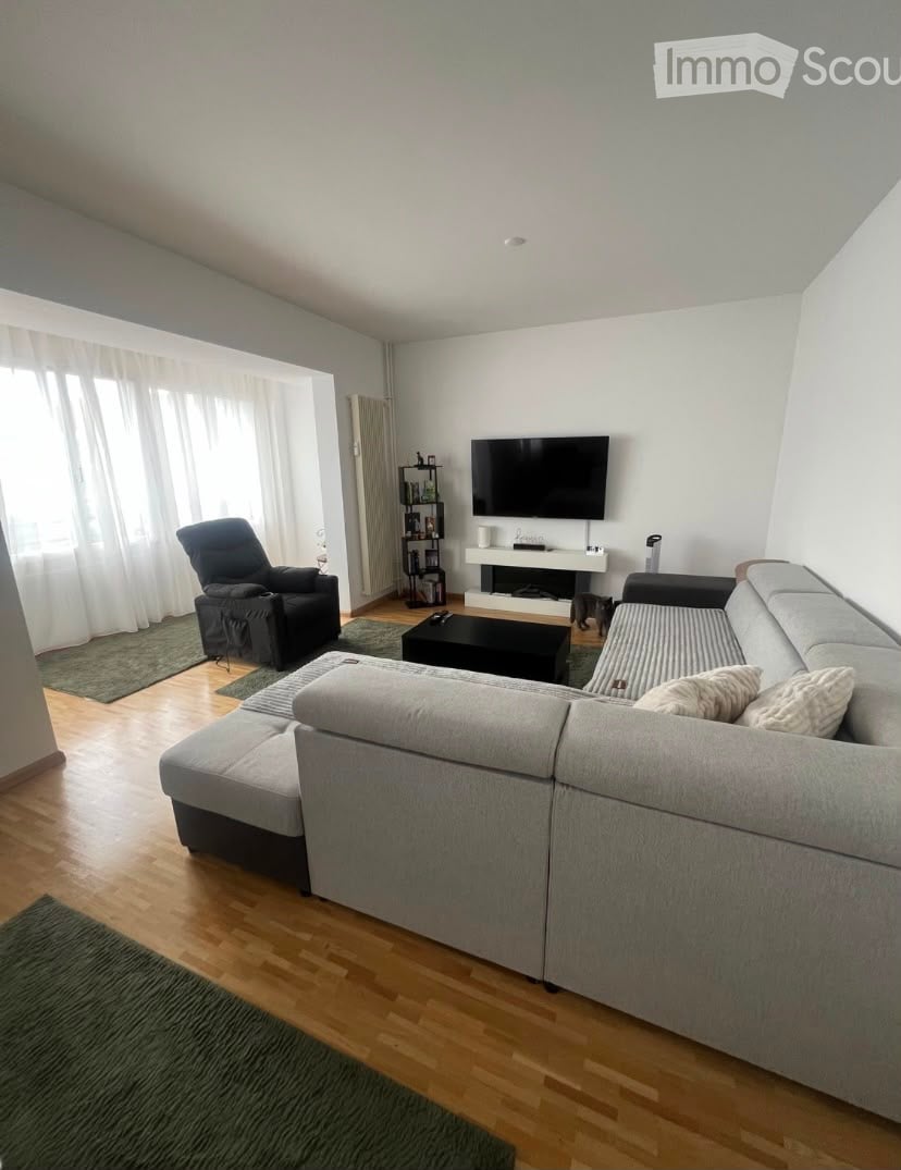 Modern living room with light grey sectional sofa, black armchair, coffee table, TV, and a shelf with decorative items.