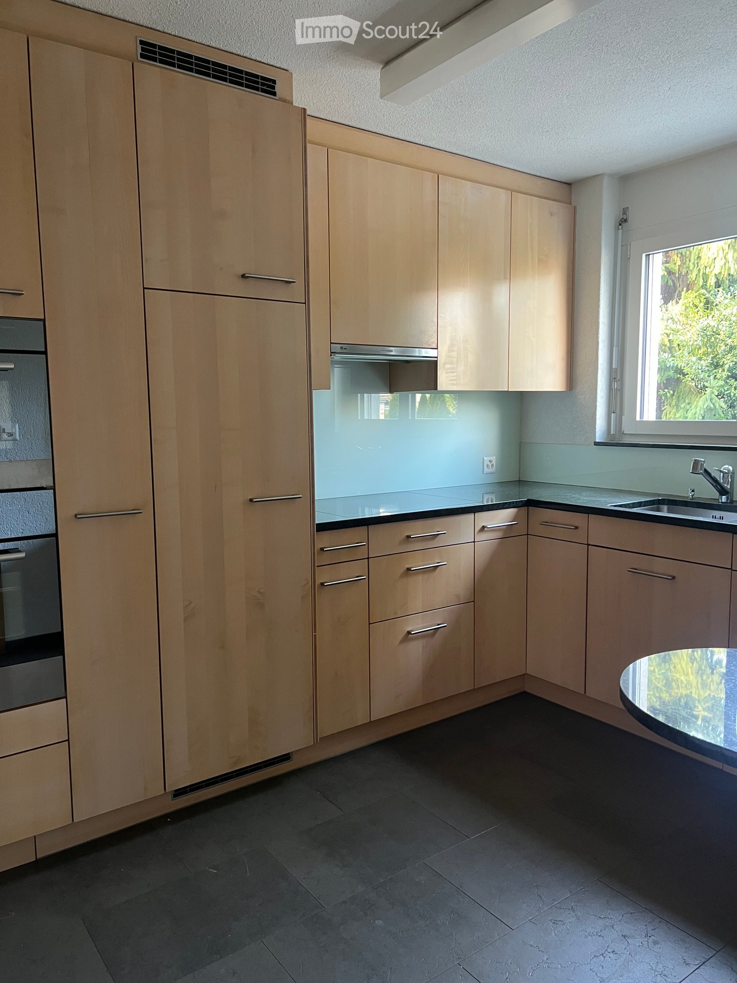 Modern kitchen with light wood cabinets, stainless steel appliances, dark tiled floor, and a small round table near the window.