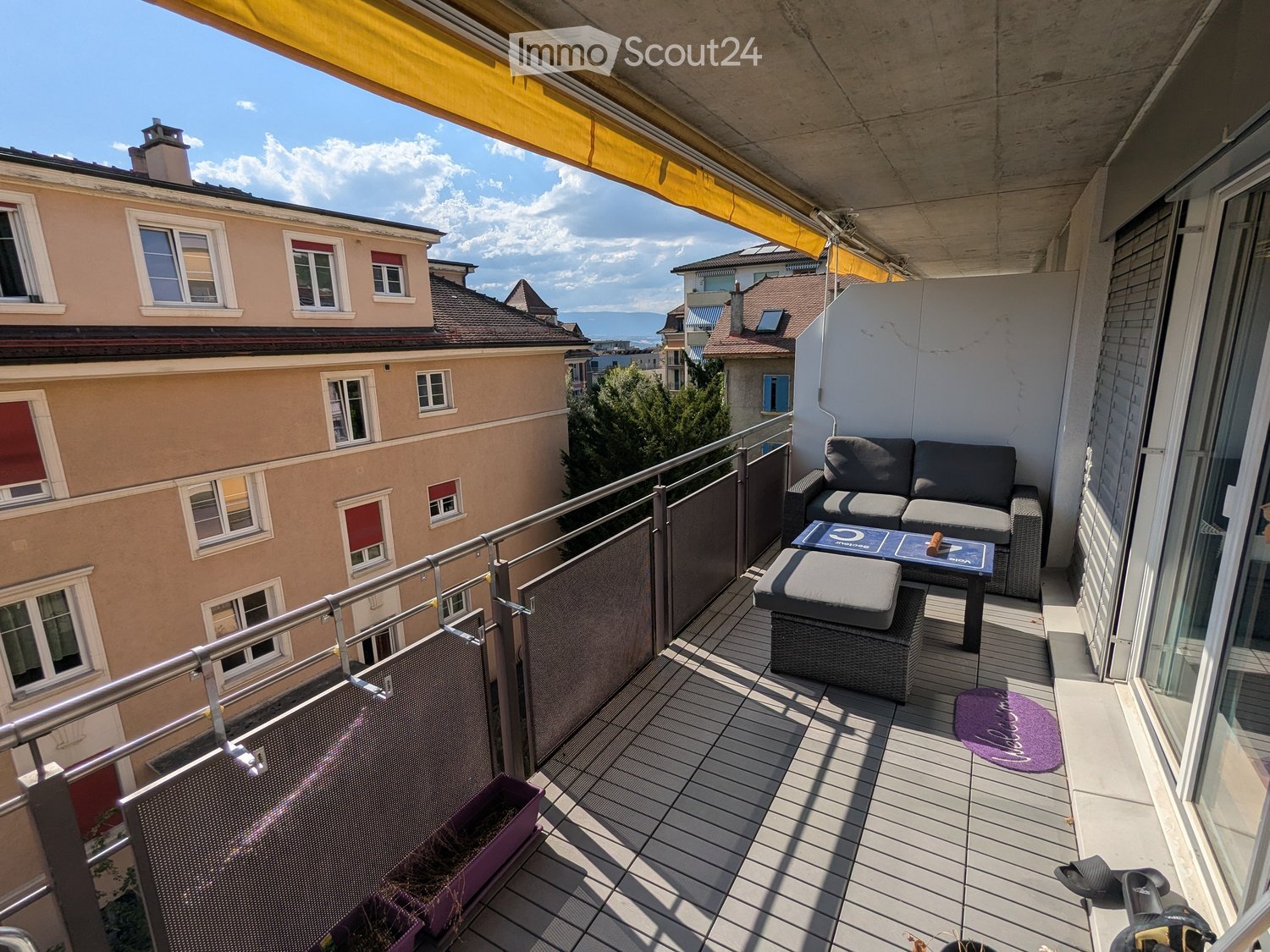 Balcony with wooden decking, metal railing, sofa, table, stool, rug, and bicycle.