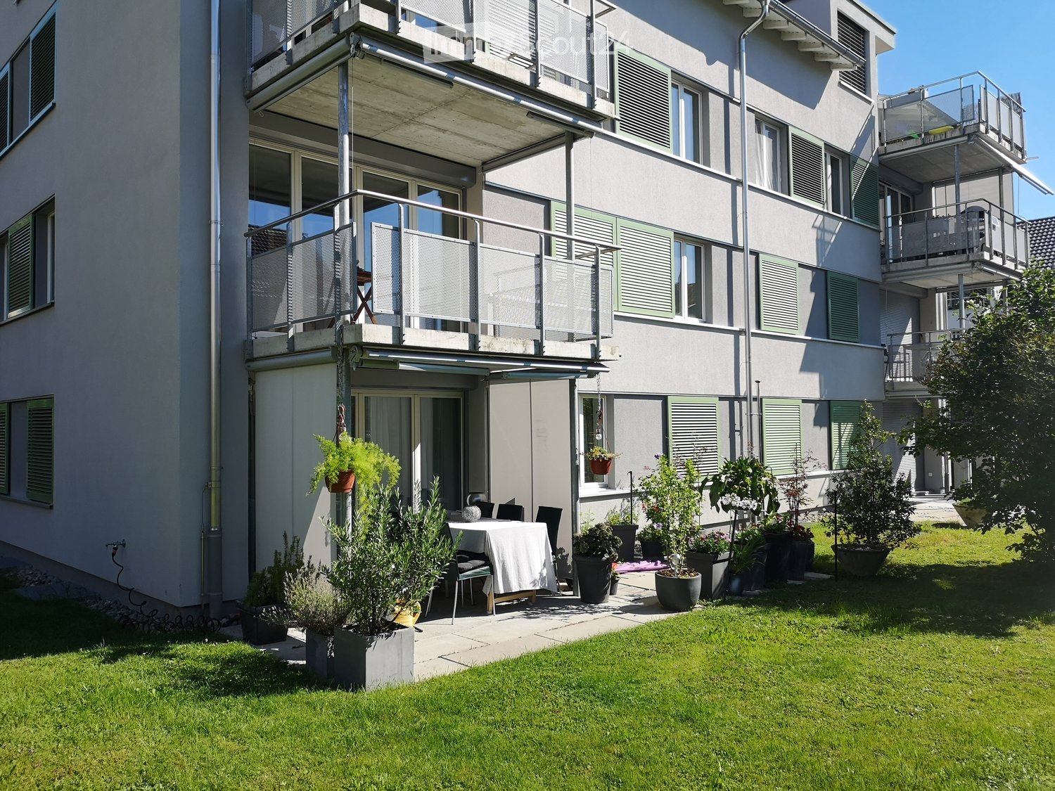 Garden area with a small patio, table, chairs, and various potted plants, adjacent to a multi-story building with balconies and windows.