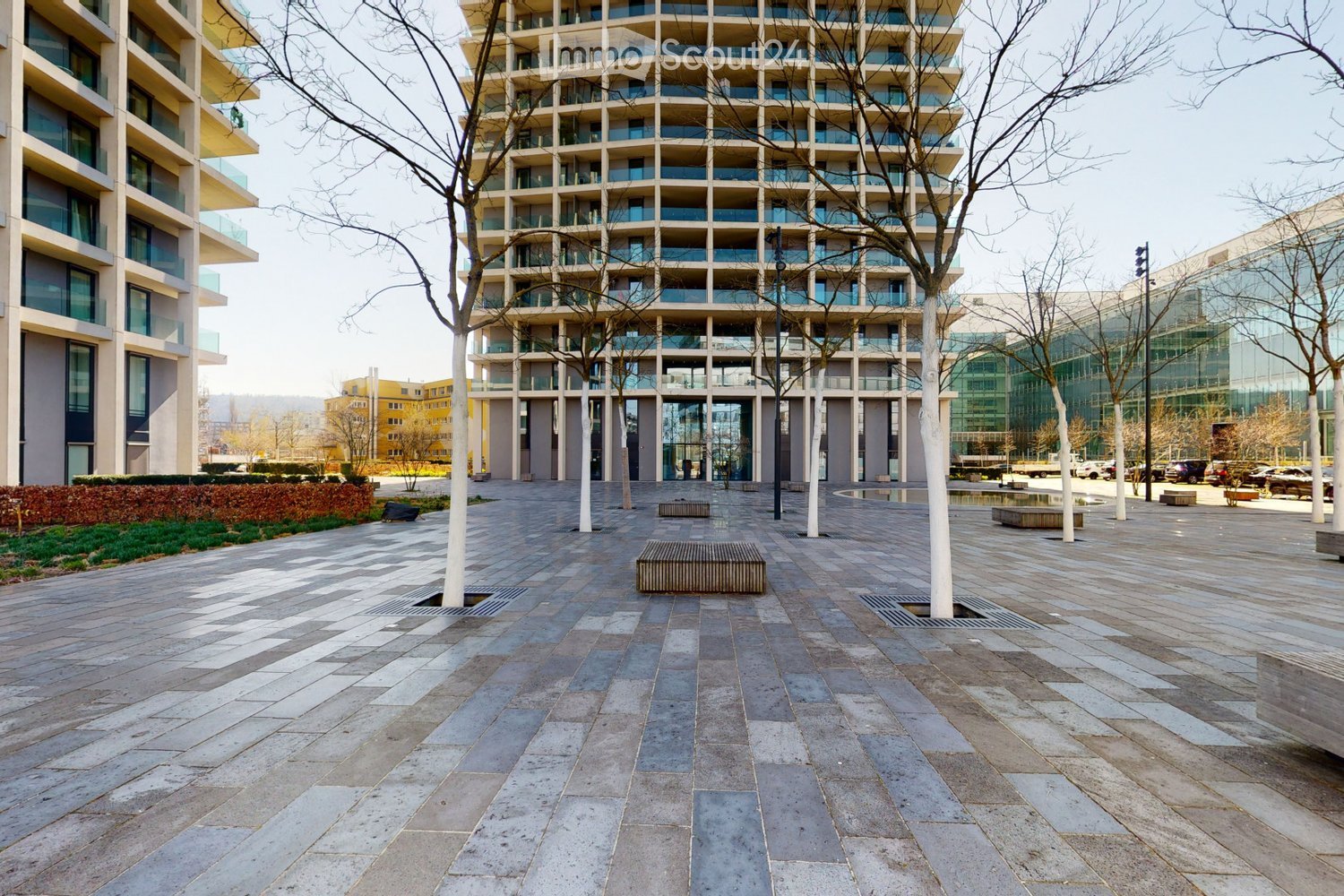Modern multi-story building with large windows and balconies, surrounded by a paved courtyard with benches and trees.