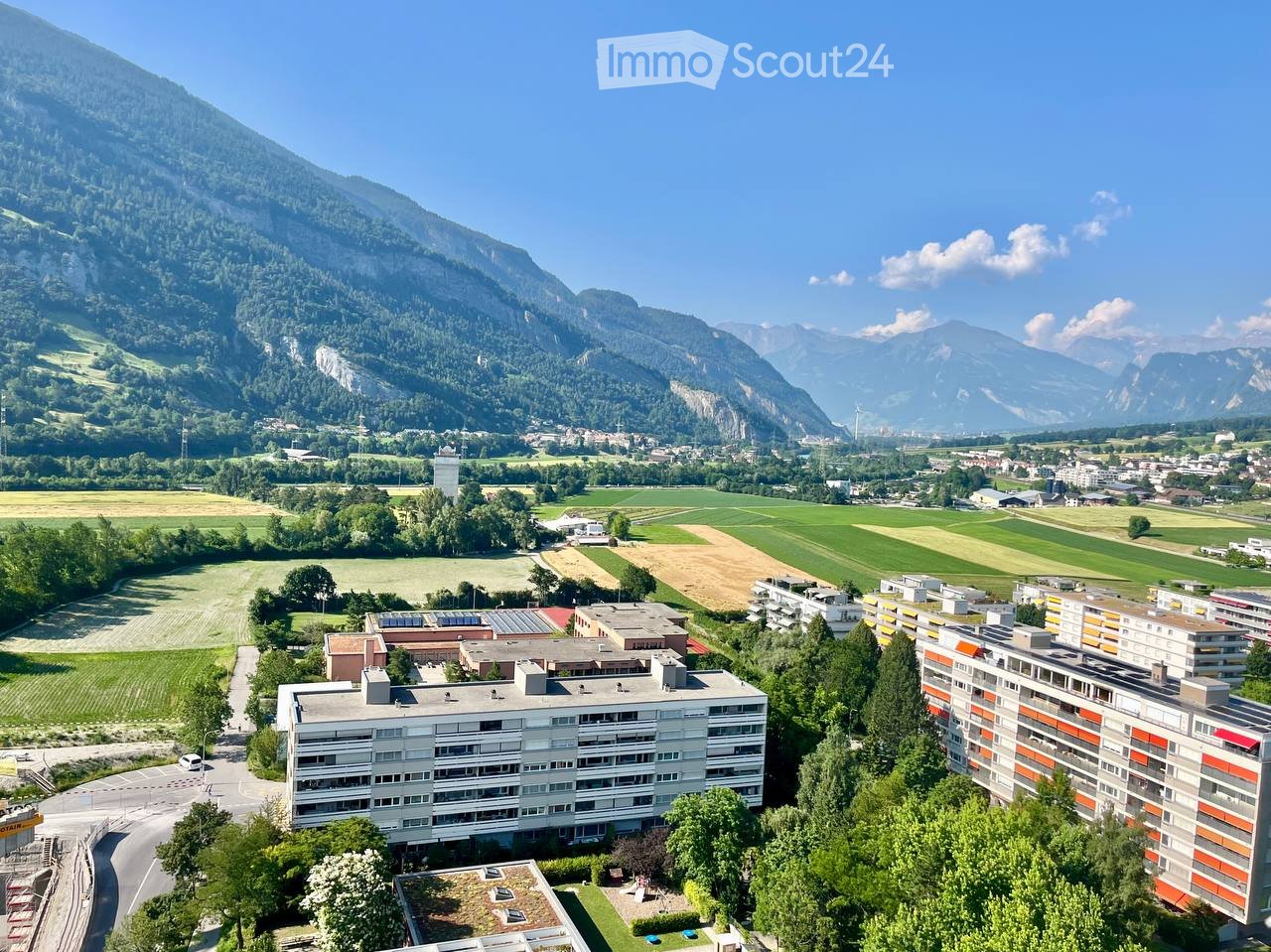 Aerial view of a residential complex surrounded by green fields and mountains in the background.