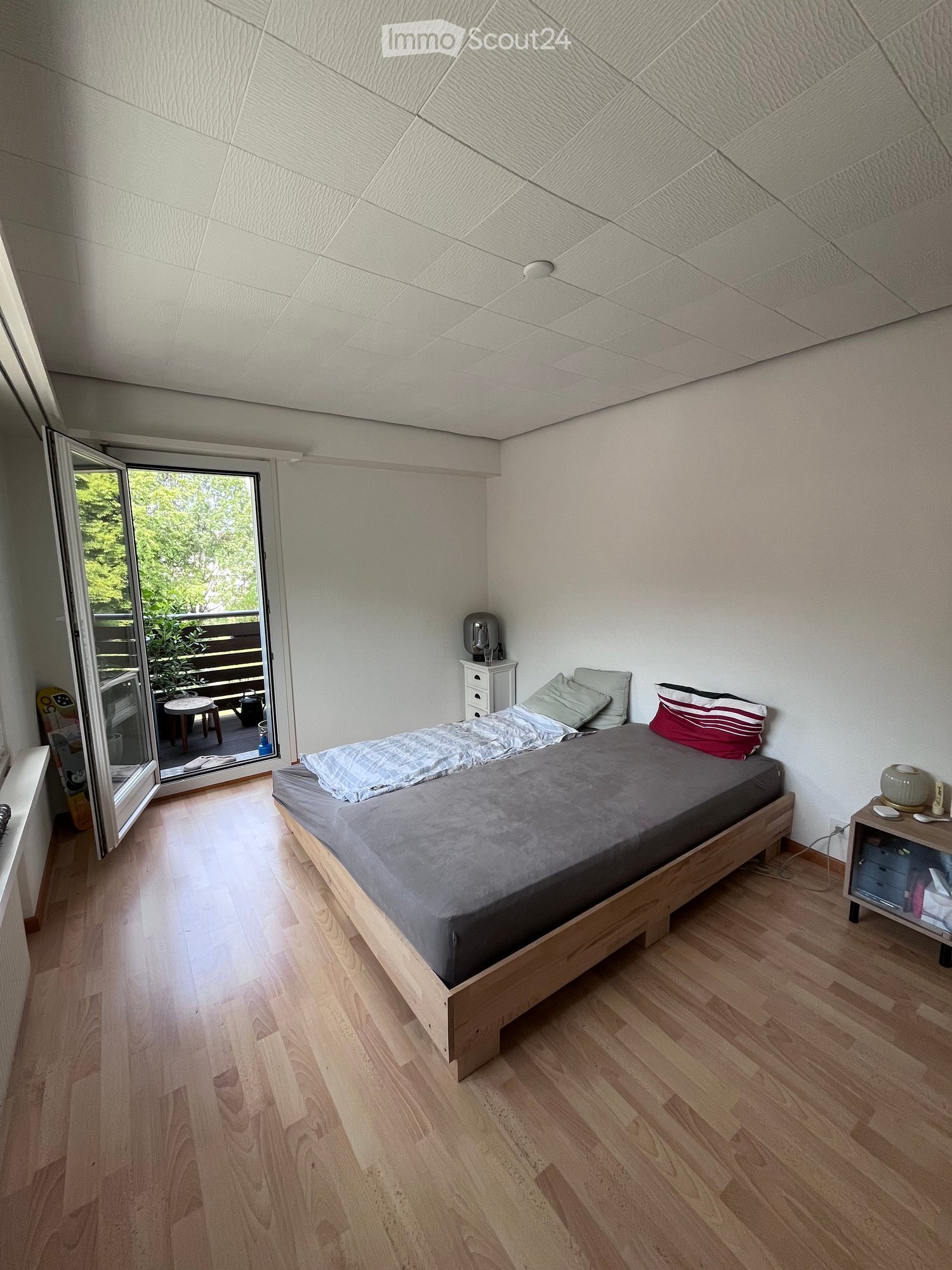 Cozy bedroom with wooden flooring, a single bed with grey bedding, nightstand, and an open sliding door leading to a balcony.
