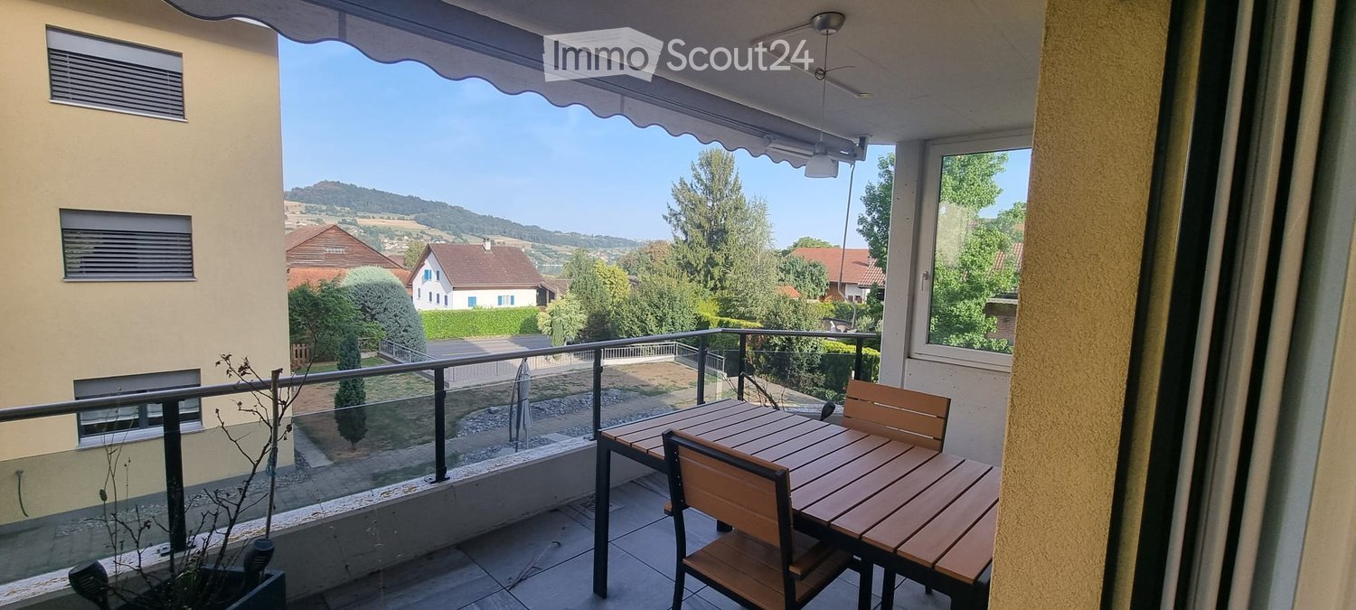 Balcony with wooden table and chairs, overlooking a scenic countryside view with houses and trees.