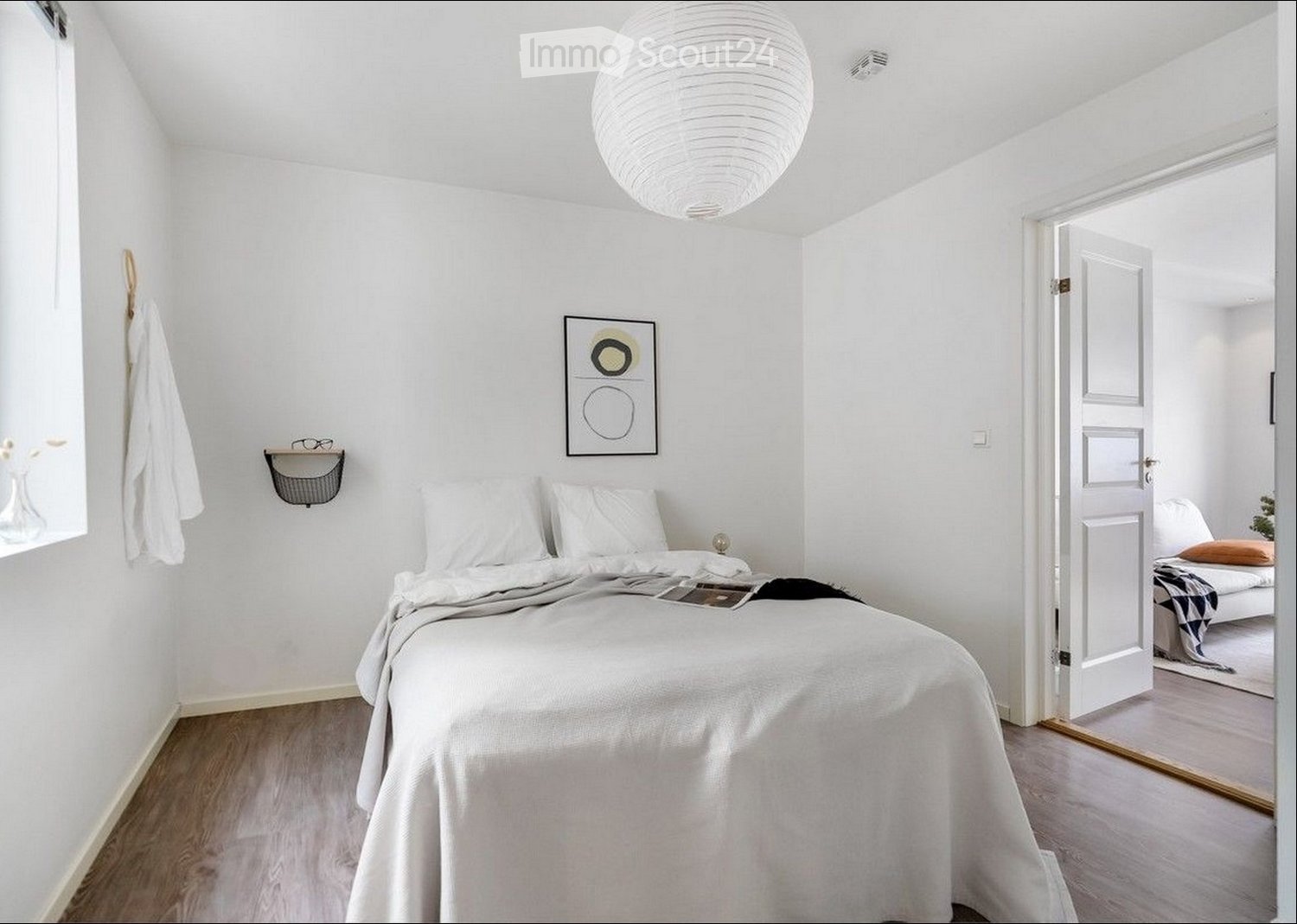 A modern, minimalist bedroom with a large bed, white bedding, a nightstand with a lamp, a picture frame on the wall, a small shelf with a basket, and a cat resting on the bed.