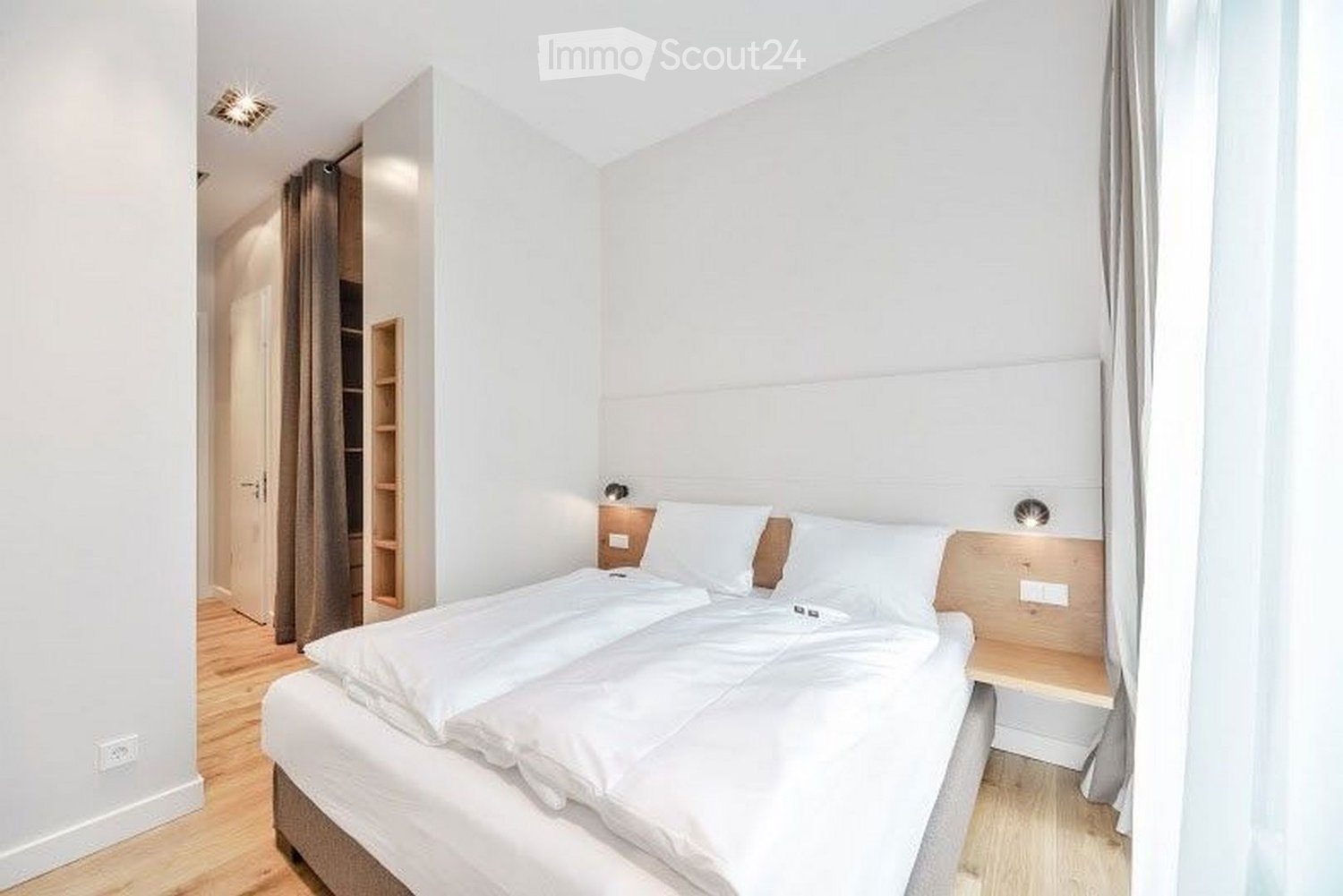 Cozy bedroom with a double bed, white bedding, a built-in nightstand with a wall-mounted light, and a curtained window. The room features light wooden flooring and white walls.