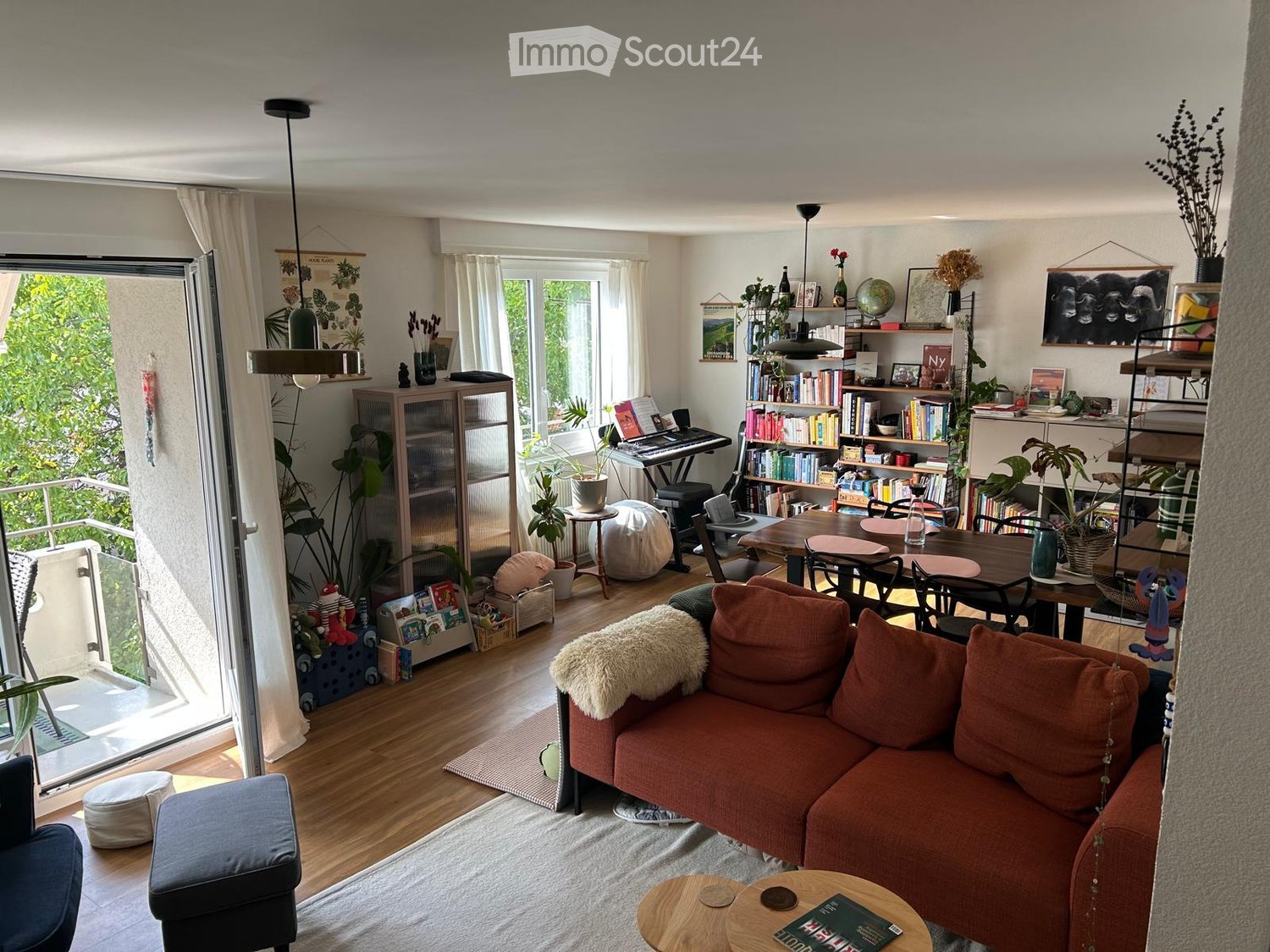 Bright living room with wooden flooring, large windows, balcony access, sofa, ottoman, small table, bookshelf, piano, and decorative plants.