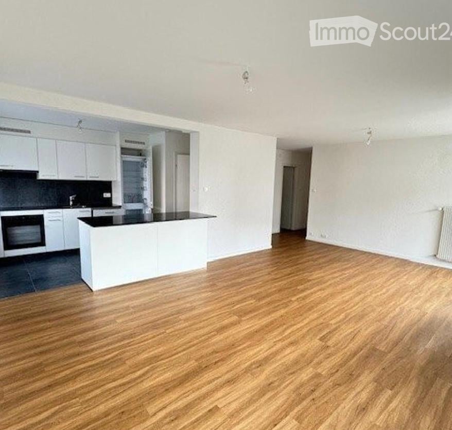 Empty room with wooden flooring, white walls, and a white kitchen area with cabinets and a countertop.