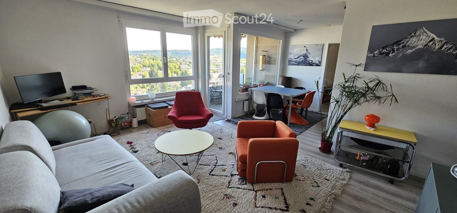 Modern living room with large windows offering a scenic view, featuring a grey sofa, round coffee table, orange armchair, dining table with chairs, and a plant in the corner.