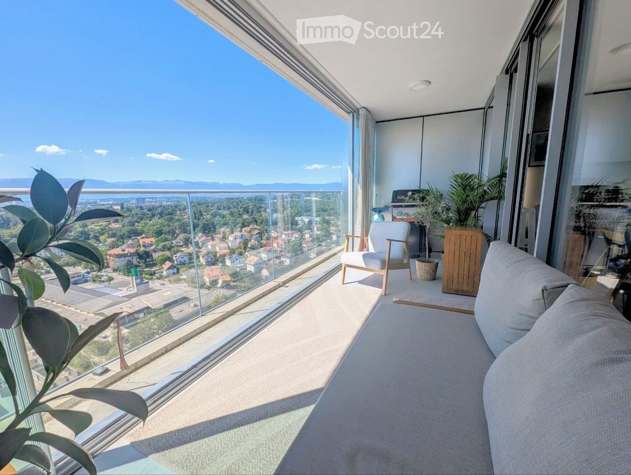 Modern balcony with large glass windows, offering a panoramic city view. Features a grey sofa, wooden chair, small table, and potted plants.