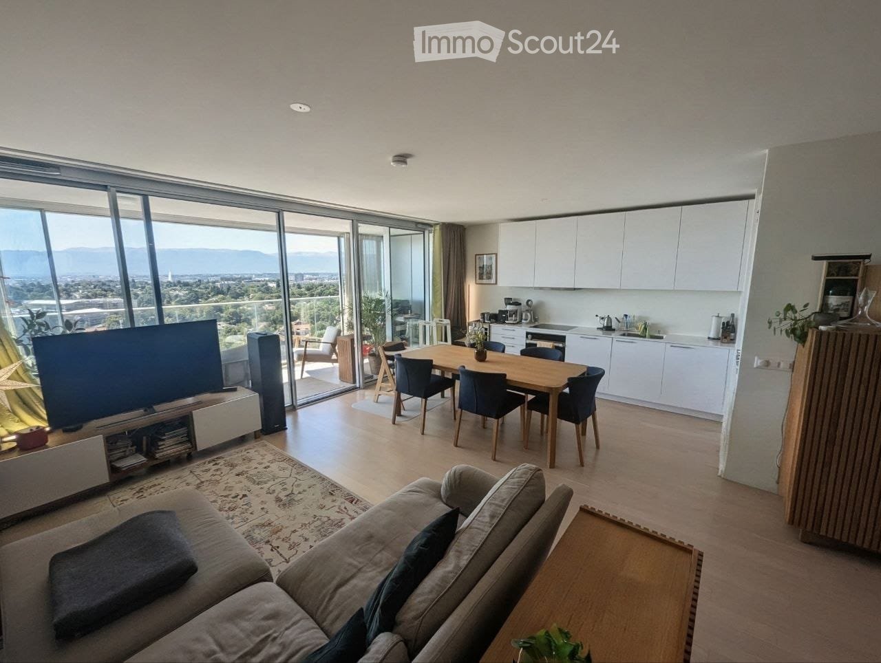 Modern open-plan living and dining area with large windows offering a scenic view, featuring a grey sofa, coffee table, dining table with chairs, and a fully equipped kitchen with white cabinets and stainless steel appliances.