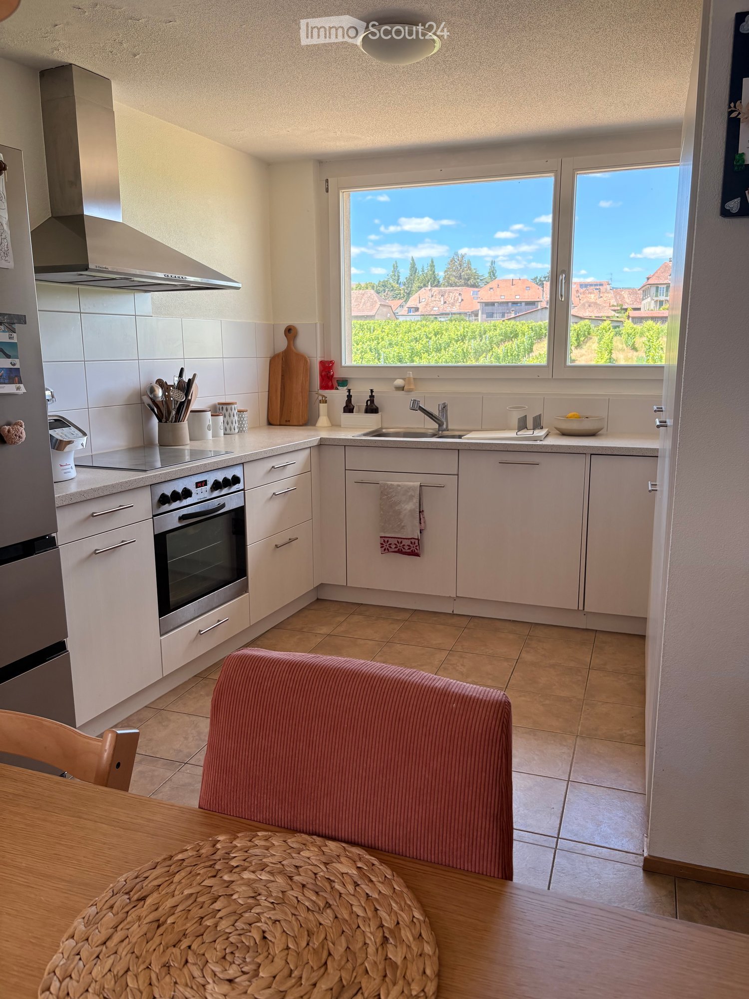 Bright kitchen with white cabinets, stainless steel appliances, and a large window overlooking a green landscape. Includes a dining area with a wooden table, woven chair, and pink bench.