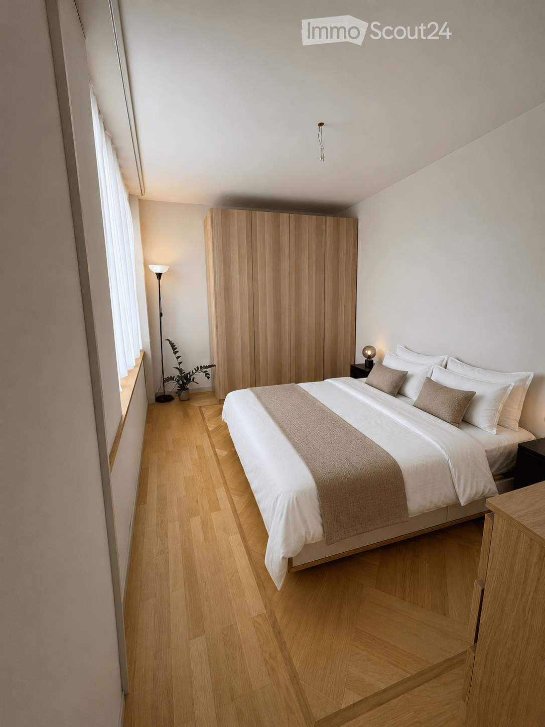 Modern bedroom with a large bed, white bedding, wooden wardrobe, nightstand, floor lamp, and a small plant near the window.