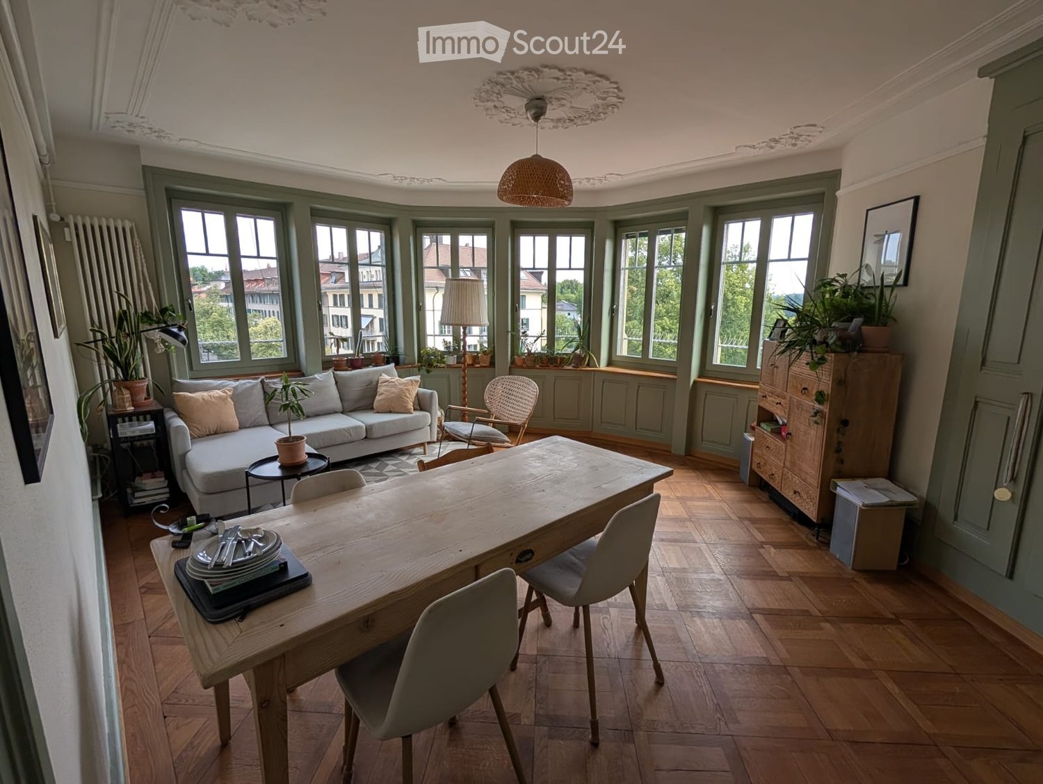 Bright living and dining area with large windows, wooden flooring, a grey sofa, a wooden dining table with chairs, and decorative plants.