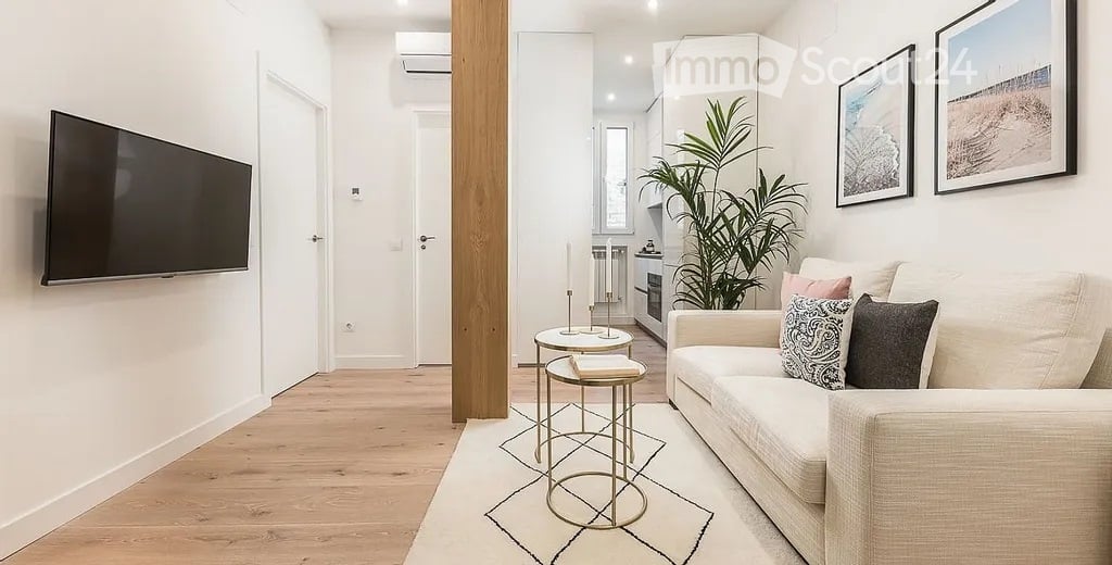 Modern living room with light-colored sofa, coffee table, small round stools, a potted plant, and a flat-screen TV mounted on the wall. The space features light wood flooring and white walls.