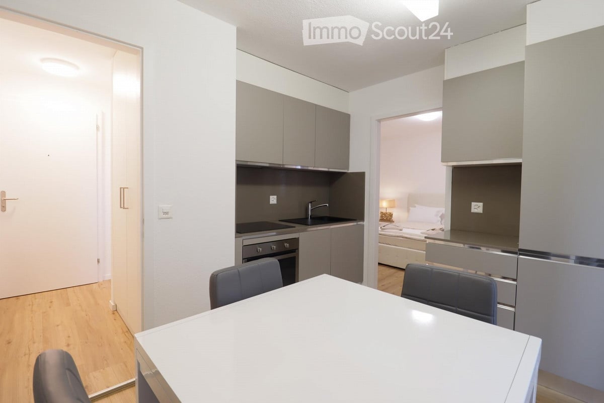 Modern, compact studio apartment with a combined living, dining, and sleeping area. Features a white dining table with chairs, a small kitchen area with cabinets, sink, and oven, and a bed with a lamp in the sleeping area.