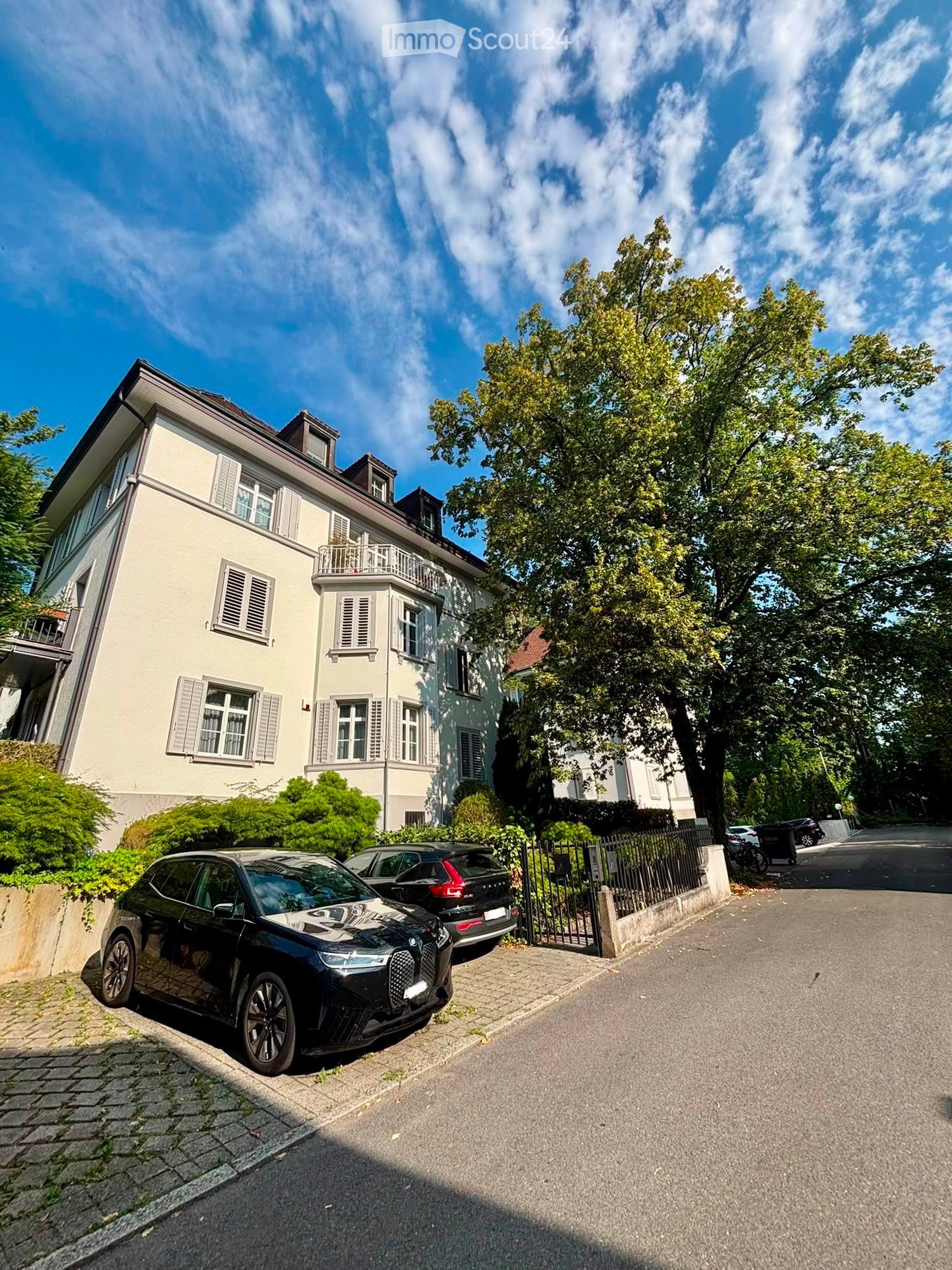 A multi-story residential house with light-colored walls, multiple windows with shutters, and a small balcony on the upper floor. The house is surrounded by greenery and has a gated driveway with parked cars.