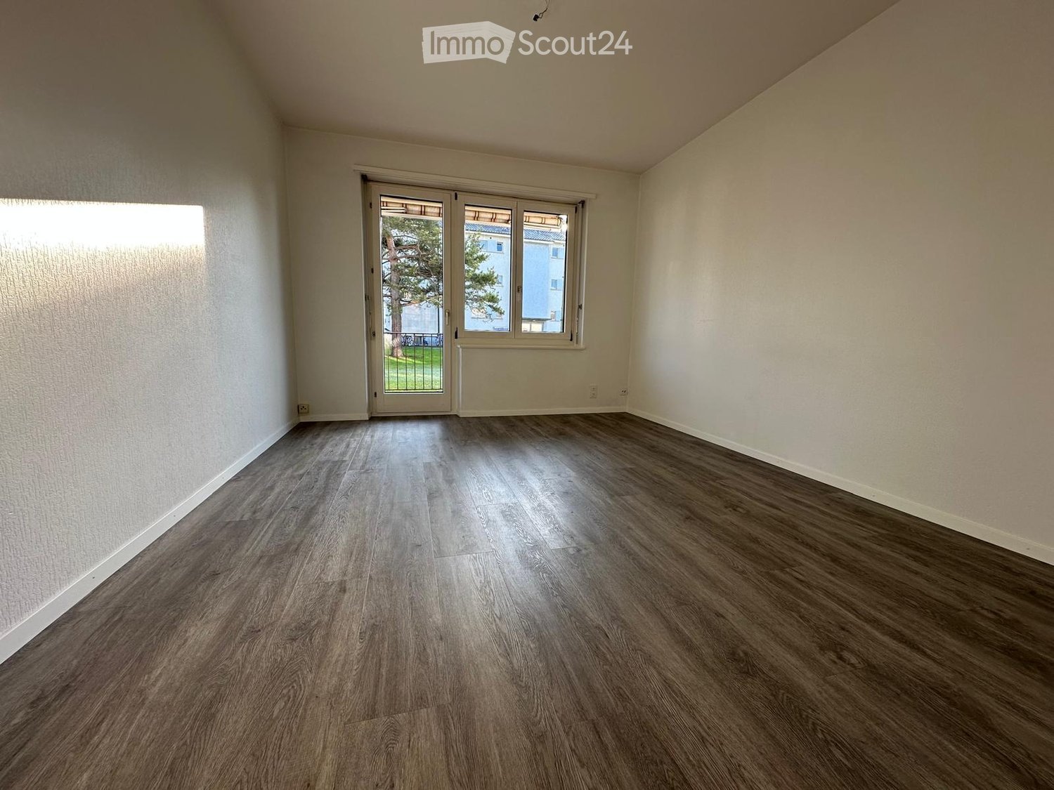 Empty room with wooden flooring, white walls, and large windows with sliding glass doors leading to an outdoor area.