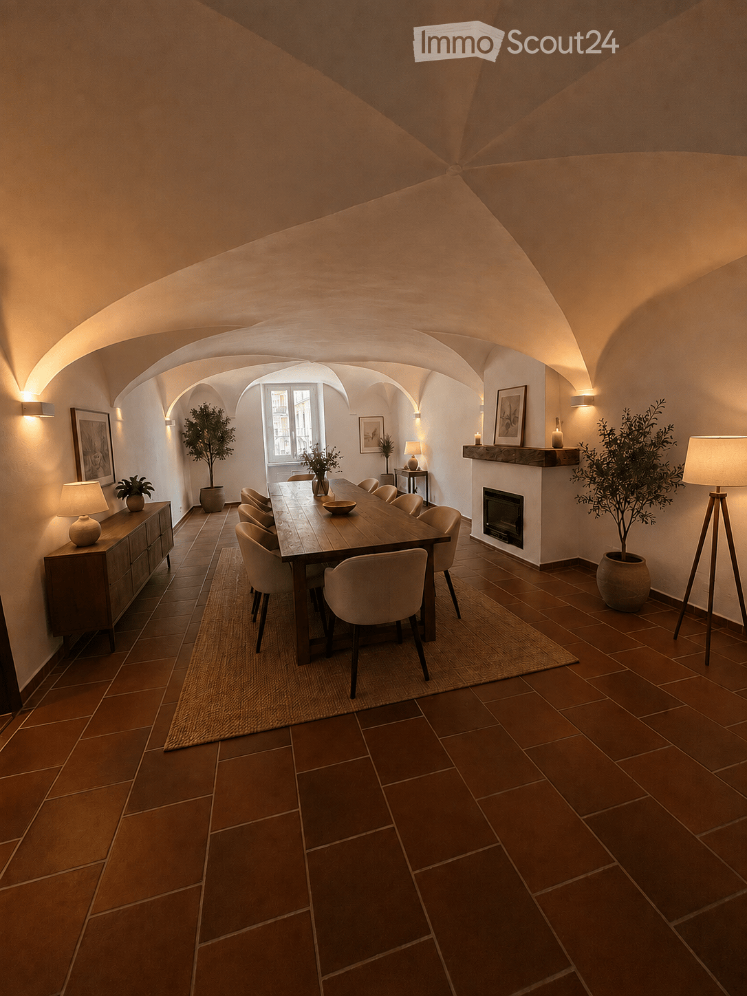 Elegant dining room with terracotta tiled floor, arched ceiling, fireplace, dining table with chairs, cabinet, lamps, and potted plants.