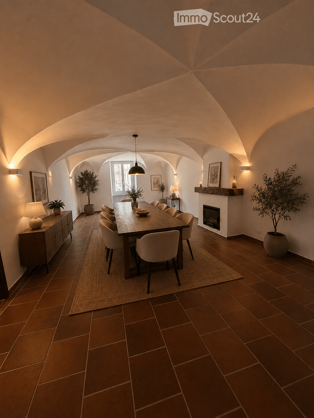 Elegant dining room with exposed wooden beams, terracotta tiles, a long wooden dining table surrounded by chairs, a fireplace, sideboard, table lamps, and potted plants.