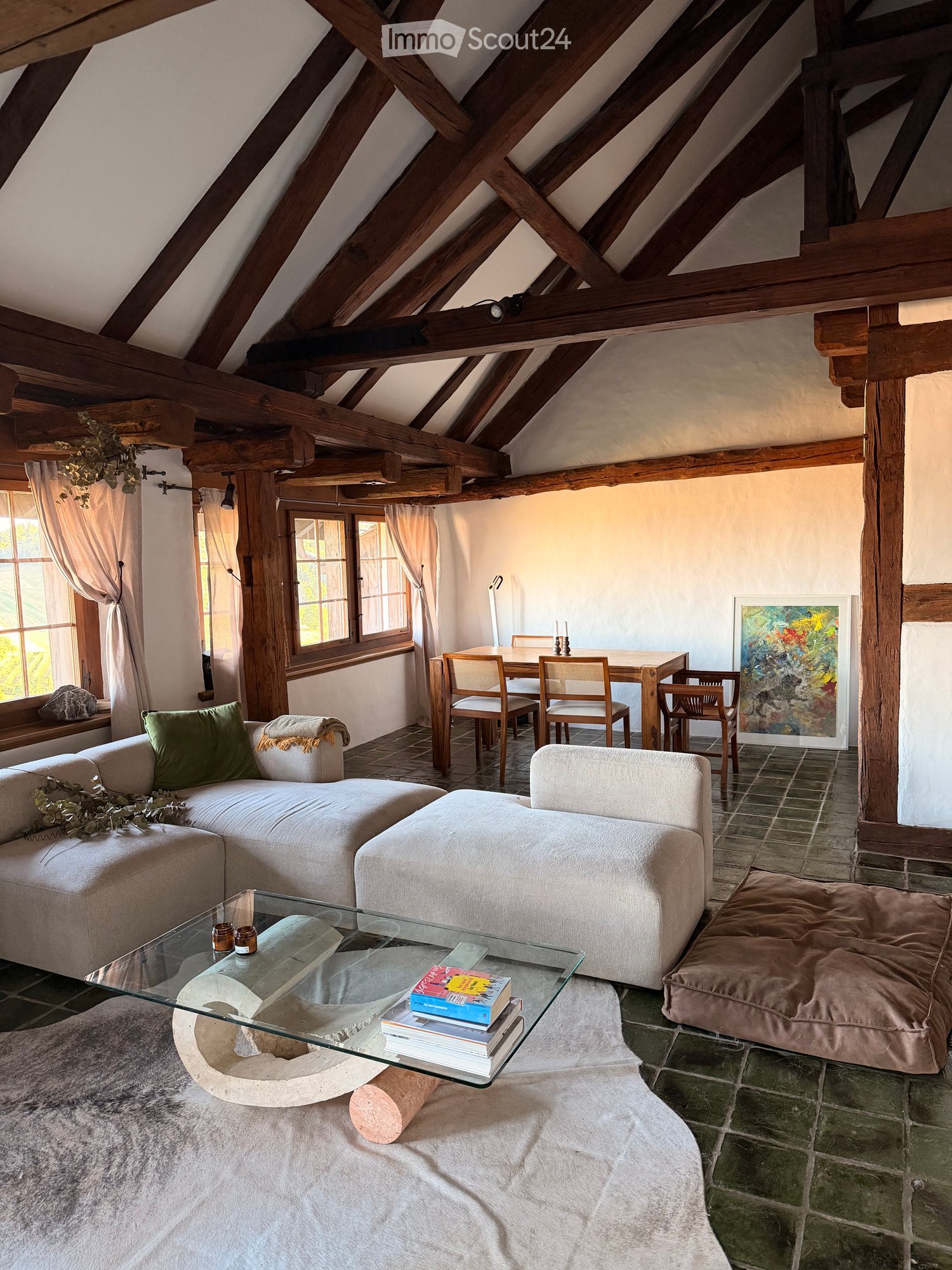 Living room with exposed wooden beams, large windows, a sofa, ottoman, coffee table, dining area with chairs and table, and a picture frame on the wall.