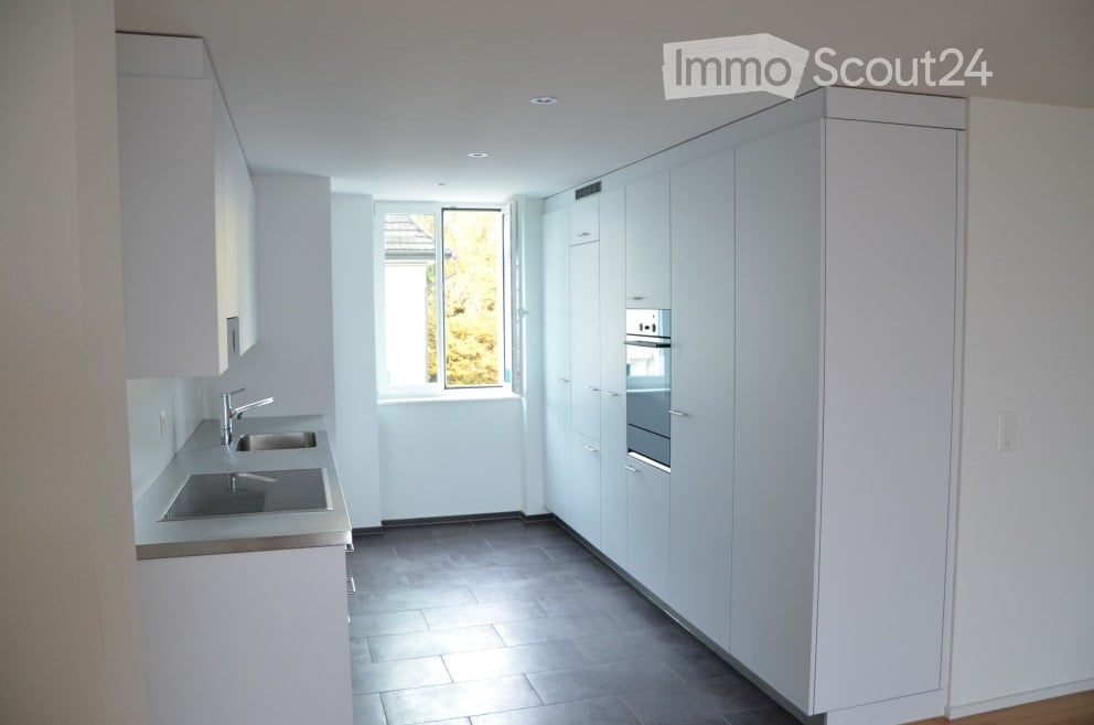 Modern, minimalist kitchen with white cabinets, stainless steel sink, and built-in oven. Features a large window with a view of greenery outside.