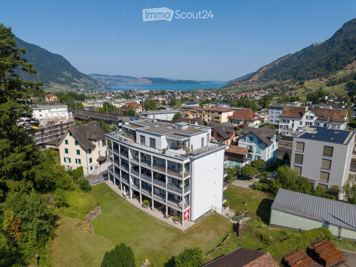 Aerial view of a modern multi-story building with balconies, surrounded by greenery and other residential structures, with a scenic lake and mountains in the background.