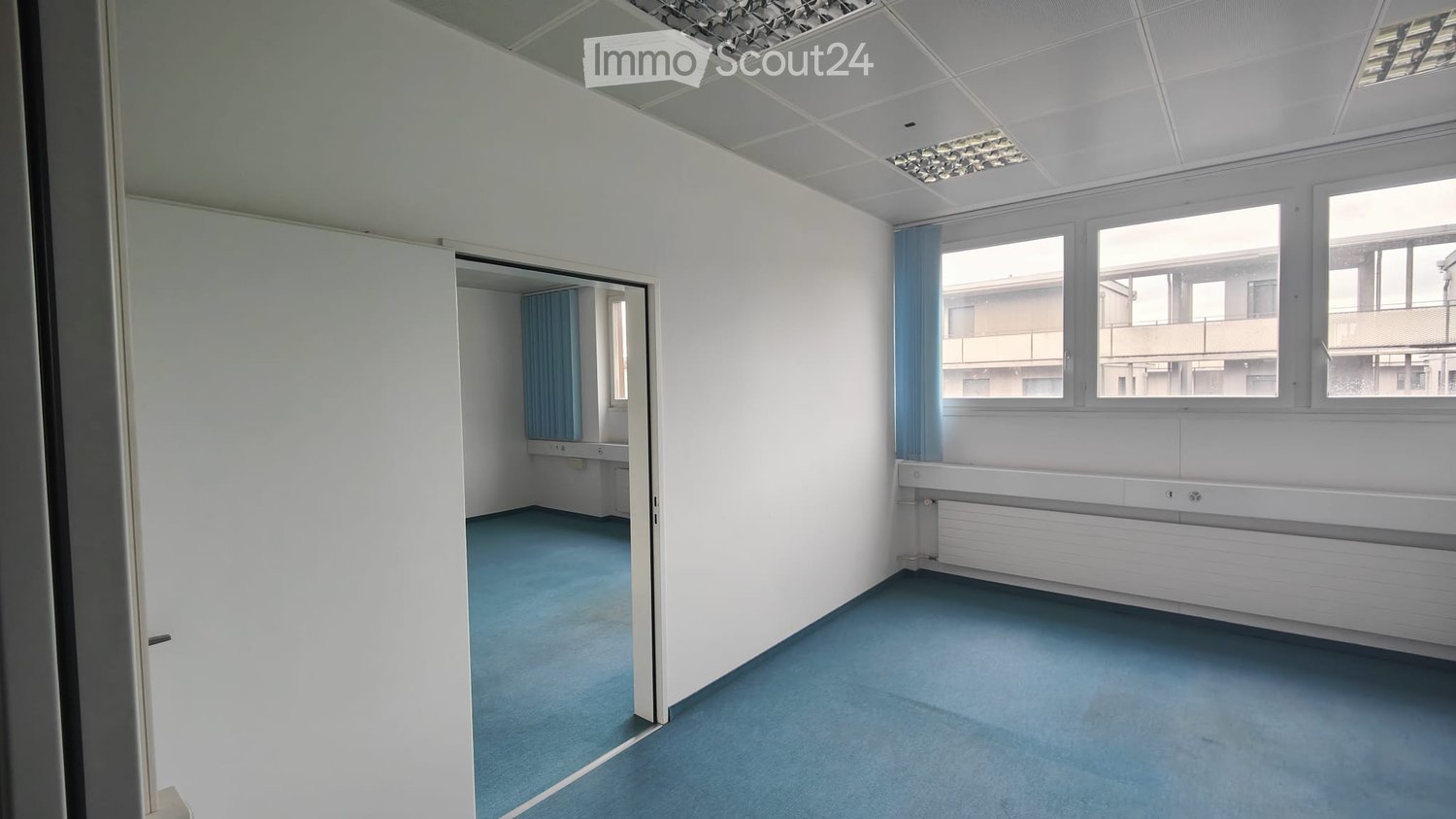 Empty room with white walls, blue carpet, and large windows with blue curtains. Adjacent smaller room visible with sink and window.