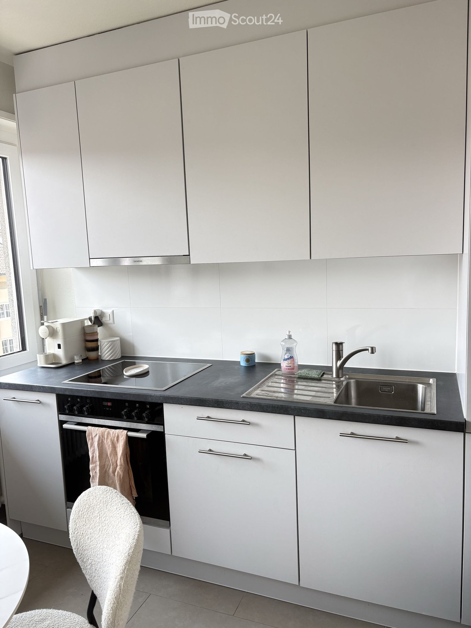 Modern kitchen with white cabinets, stainless steel appliances, and a black countertop. Features an oven, sink, faucet, and a white chair.