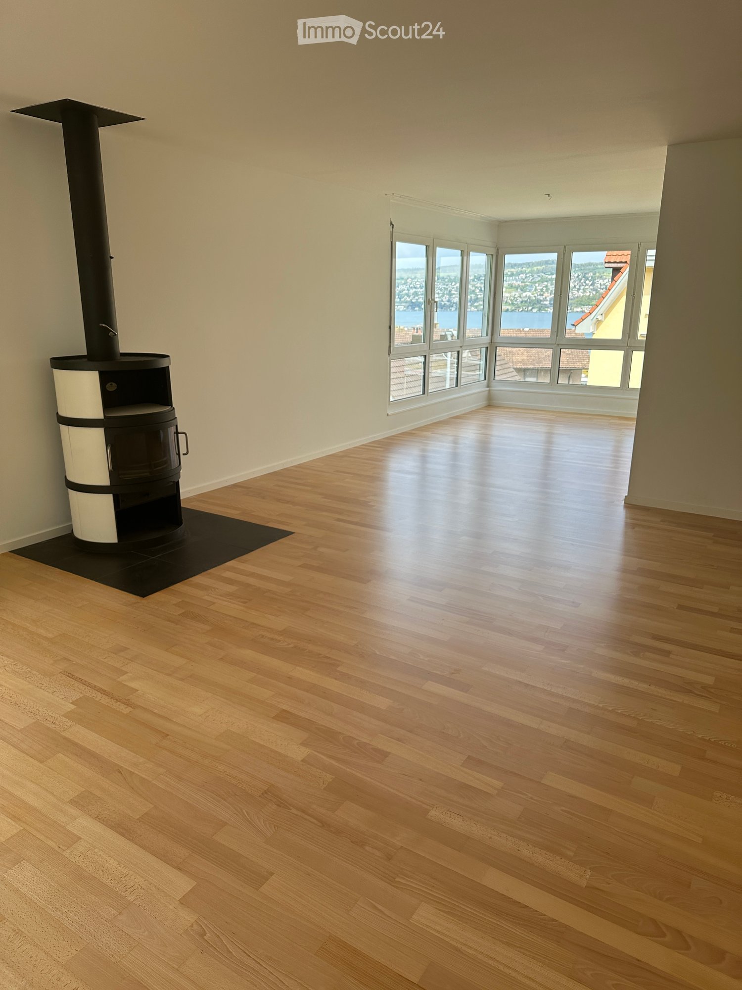 Empty room with wooden flooring, large windows offering a scenic view, and a modern black and white wood-burning stove.