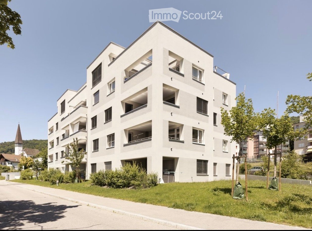 Modern multi-story residential building with white facade, numerous windows, and balconies. Surrounded by greenery and a paved pathway.