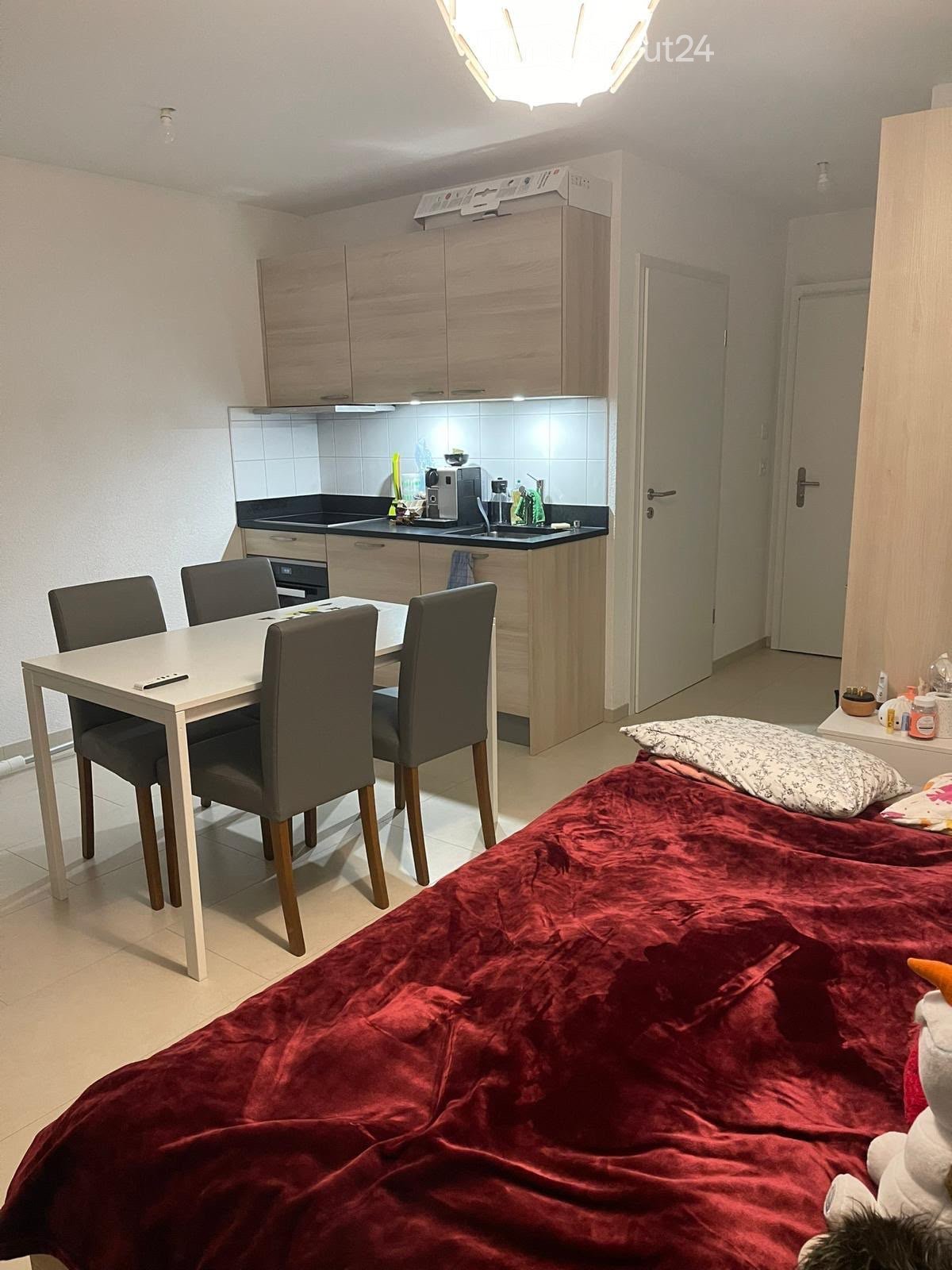 A compact studio apartment with a bed, a small dining table with chairs, and a kitchen area featuring cabinets, a sink, and a refrigerator.