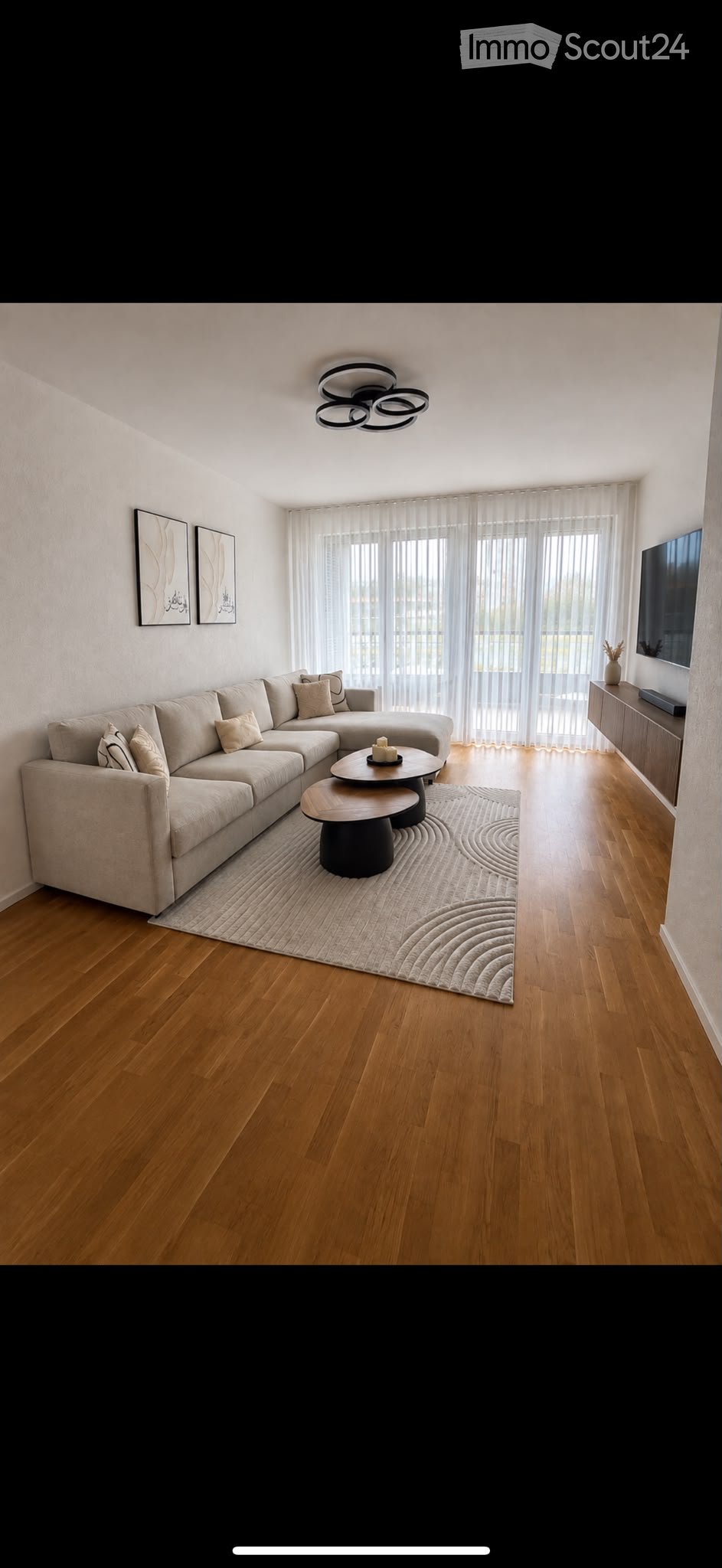 Modern living room with a sectional sofa, coffee table, TV, and large windows with sheer curtains.