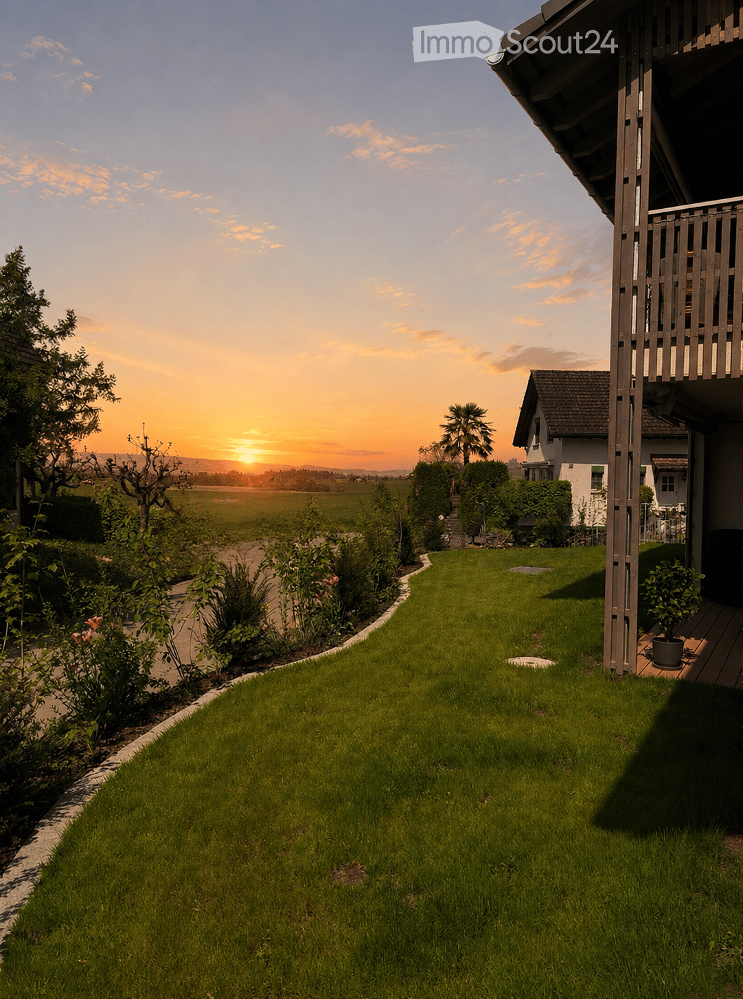 Well-maintained lawn with a curved pathway, surrounded by shrubs and flowers, leading to a house with a balcony and a view of a sunset over a field.