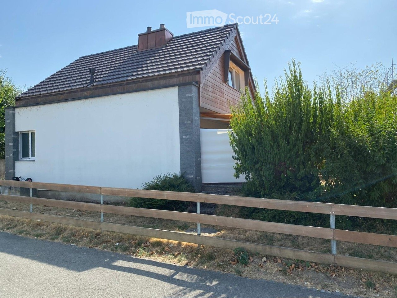 Single-story house with a pitched roof, white exterior wall, wooden facade section, chimney, and a wooden fence in front.