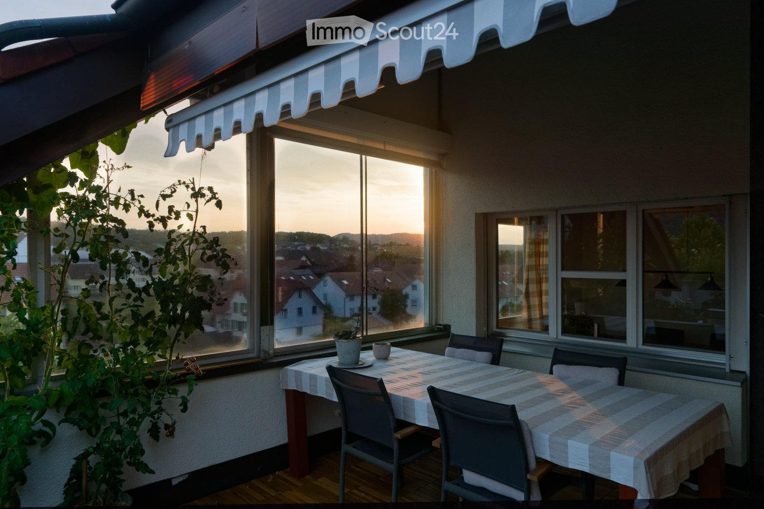 Balcony with a table, chairs, and a potted plant, overlooking a town with houses and a sunset view.