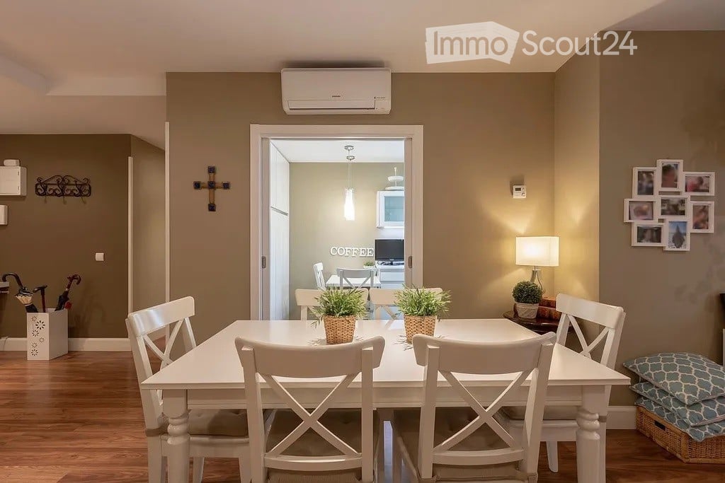 Dining room with a white dining table and chairs, a lamp on a side table, and potted plants on the table. An open doorway leads to a kitchen area with a TV and dining setup.