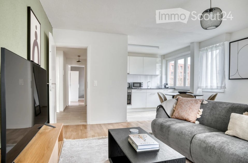 Modern living room with grey sofa, coffee table, books, and pillows. Adjacent kitchen area with white cabinets, microwave, and dining table with chairs.