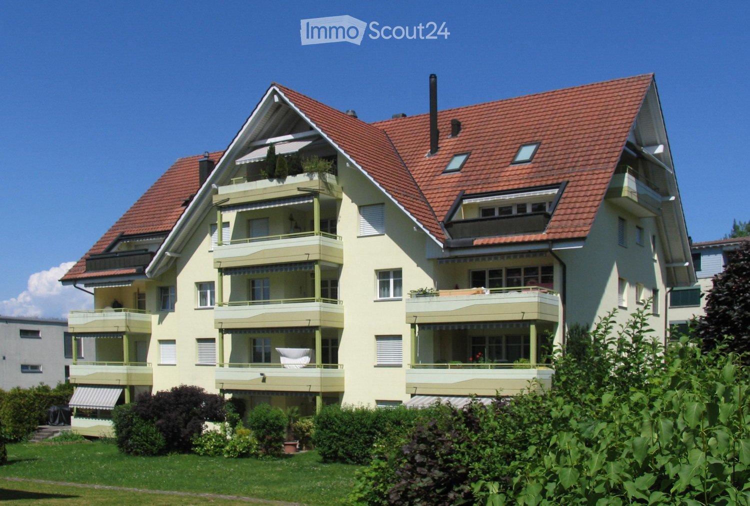 A two-story house with a red-tiled roof, multiple balconies, and green shutters. The house is surrounded by a well-maintained garden with various plants and shrubs.