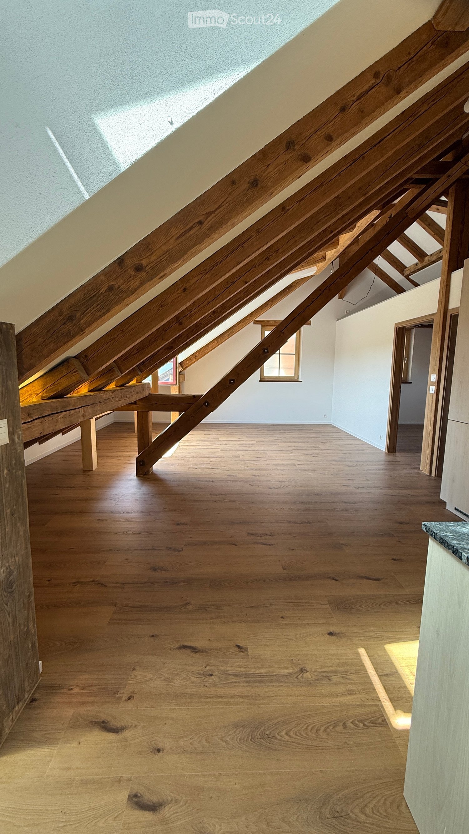 An attic space with exposed wooden beams, wooden flooring, and a skylight window allowing natural light to enter. The room is empty, with no furniture or furnishings visible.