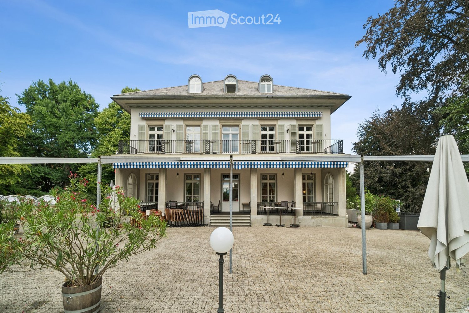 A two-story building with a balcony and outdoor seating area, surrounded by greenery and a paved courtyard.