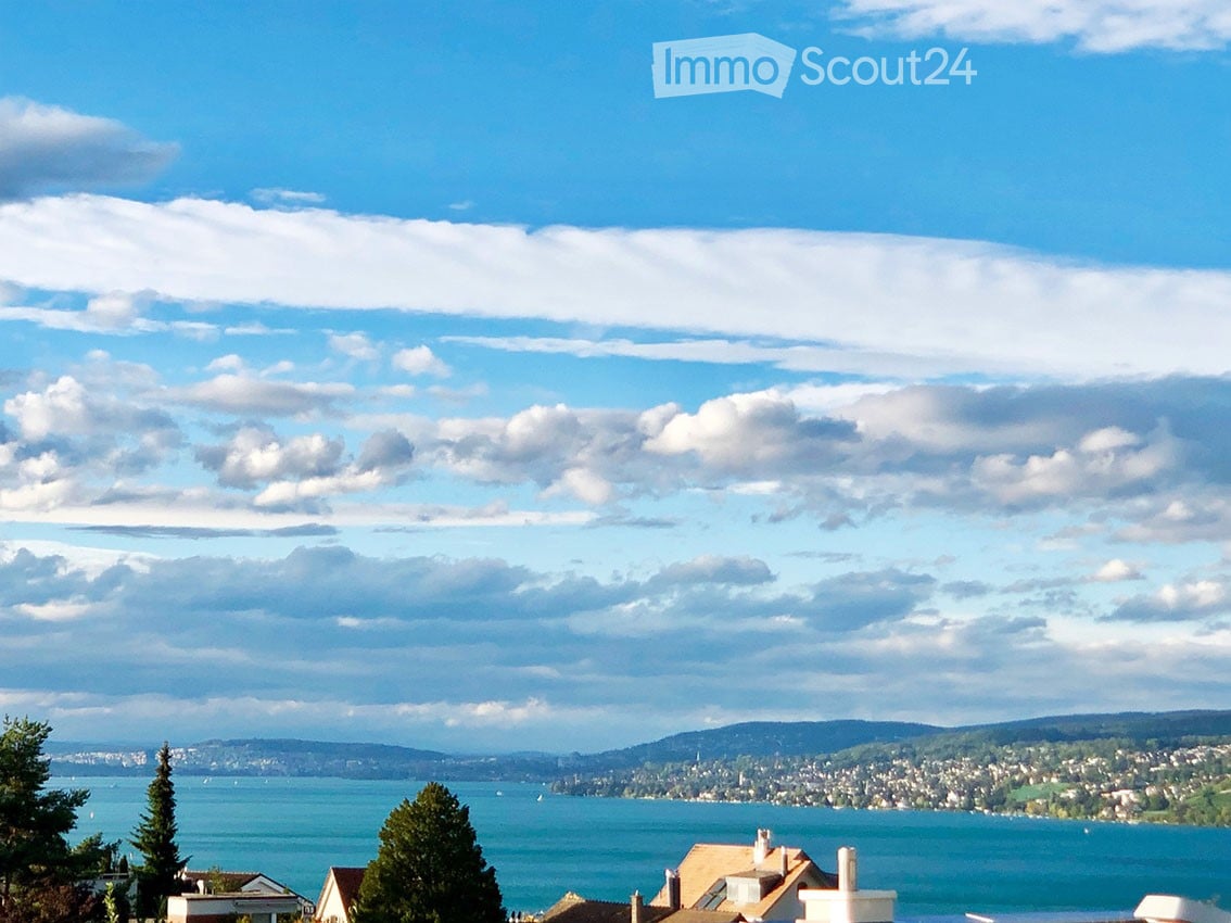 Scenic view of a lake with surrounding hills and a town in the distance under a partly cloudy sky.
