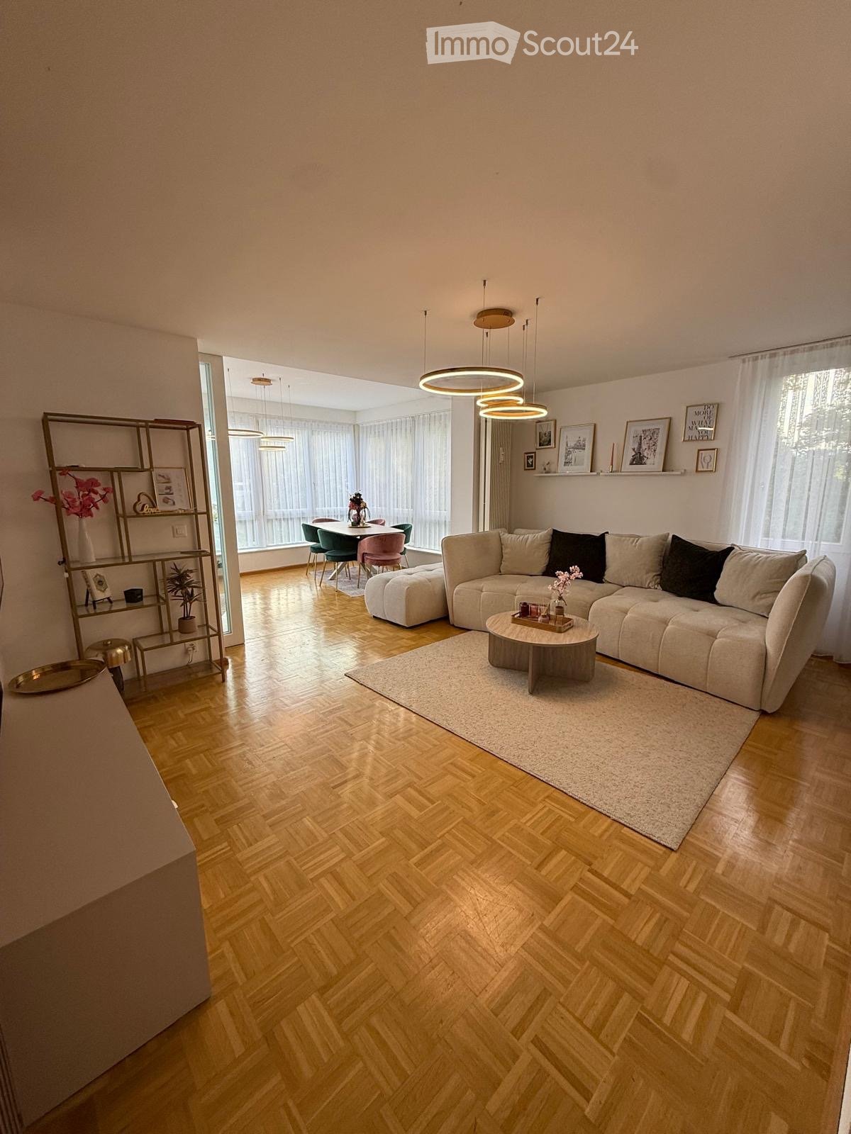 Modern living room with light wooden herringbone flooring, large windows with white curtains, a sectional sofa, coffee table, dining area with chairs and table, and a decorative shelf with vases and picture frames.