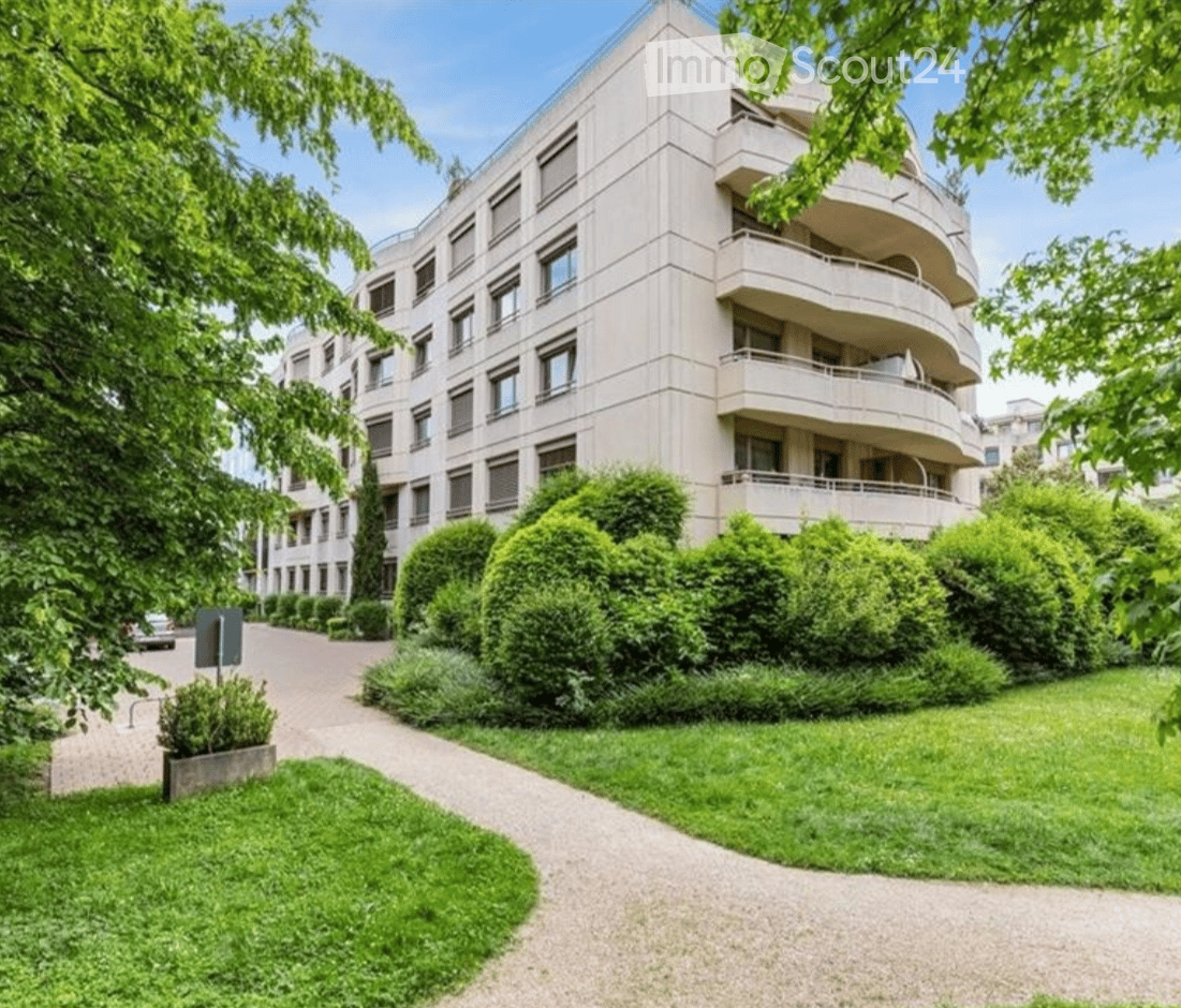Modern multi-story apartment building with balconies, surrounded by well-maintained greenery and a paved pathway.
